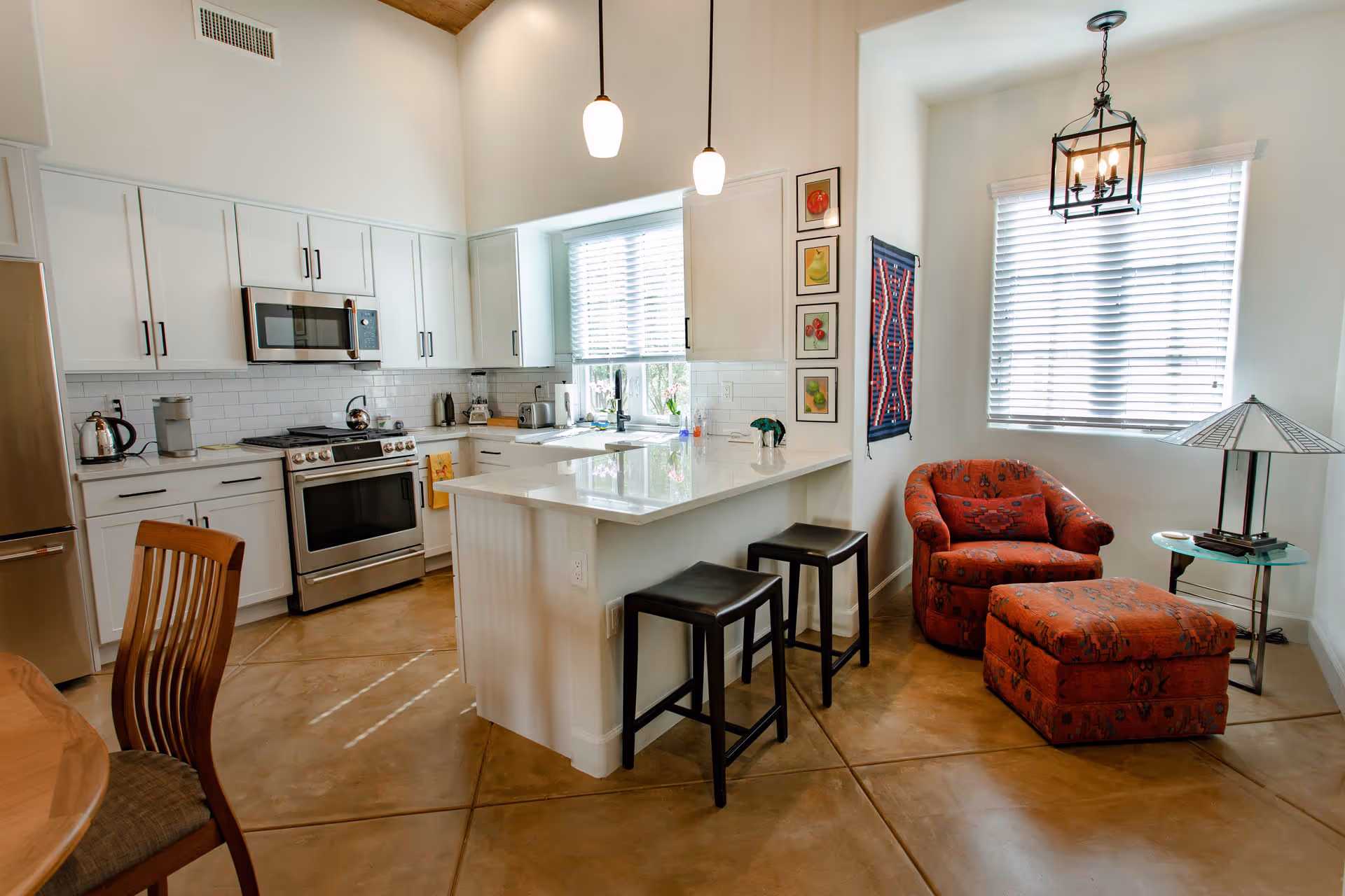 Bright kitchen and small sitting area with white cabinets, stainless steel appliances, a kitchen island with two black stools, a red patterned armchair with matching ottoman, a small glass side table with a lamp, and two windows with blinds.
