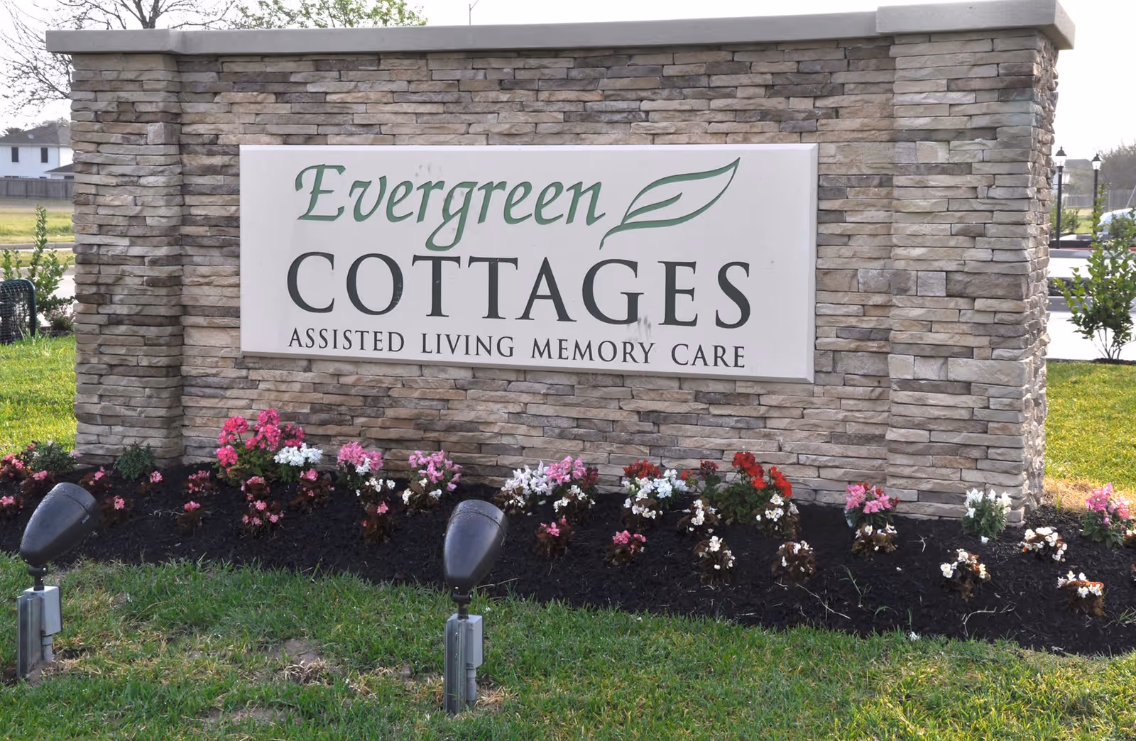 Stone sign for Evergreen Cottages Assisted Living Memory Care with flowers planted in front and grass surrounding it.