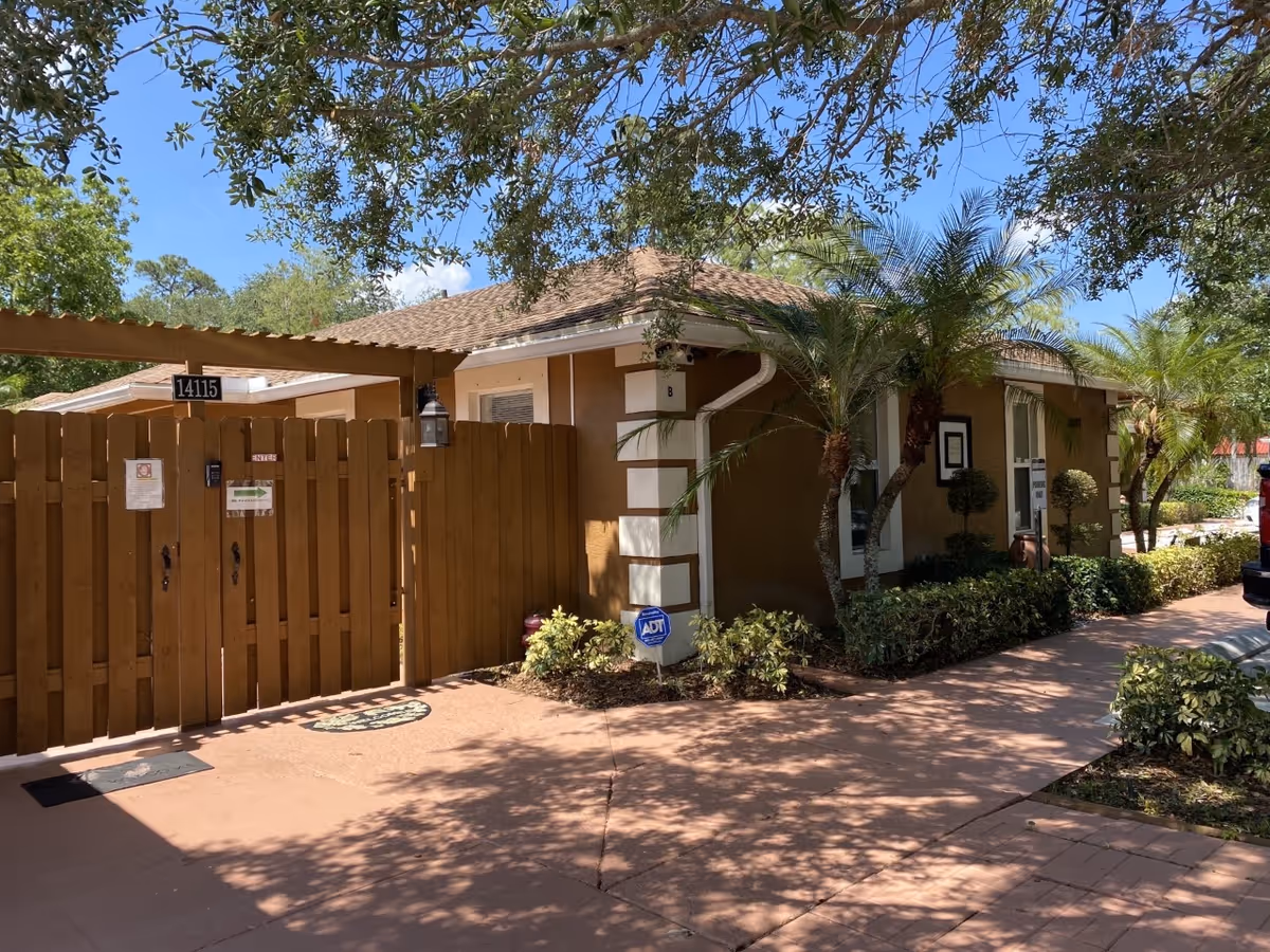 Front entrance of a single-story assisted living building with a wooden gate, palm trees, and a paved walkway.