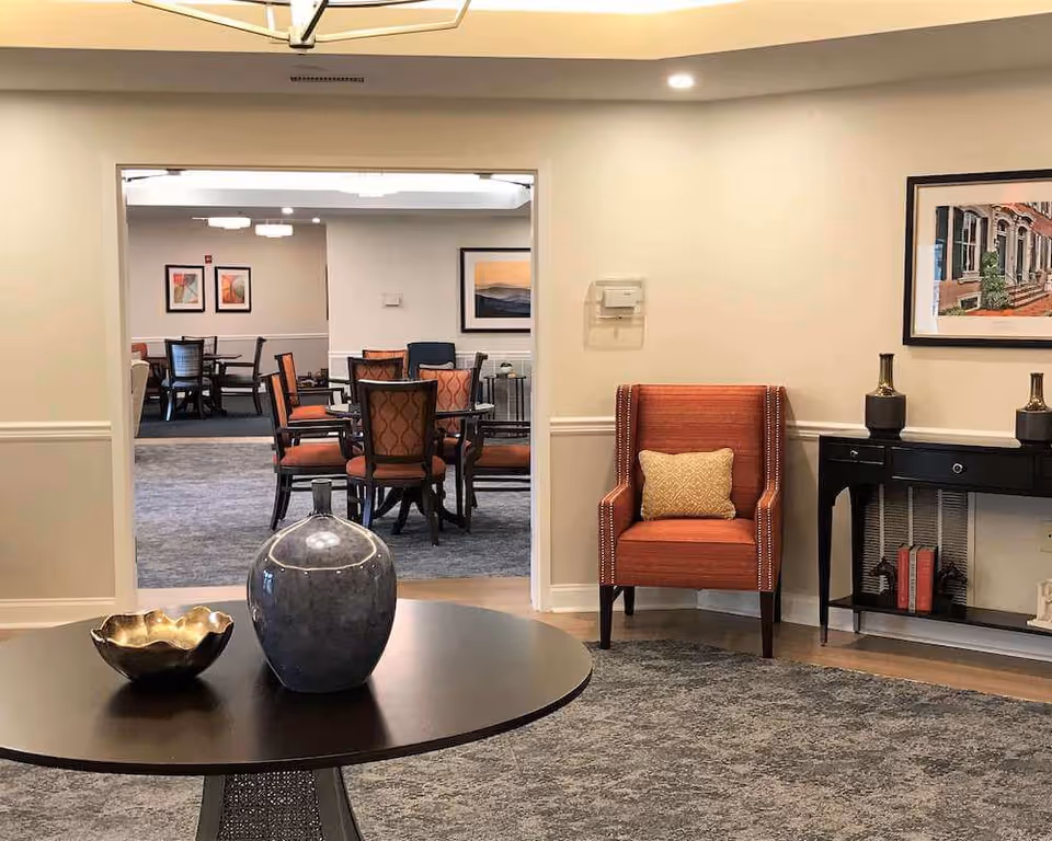 A cozy interior space in Arbor Terrace Herndon featuring a round table with decorative items including a large blue vase and a gold bowl. To the right, there is an orange armchair with a beige cushion next to a black console table with two decorative vases and books underneath. The wall has framed artwork. Through an open doorway, a dining area with multiple tables and chairs is visible, decorated with framed pictures on the walls.