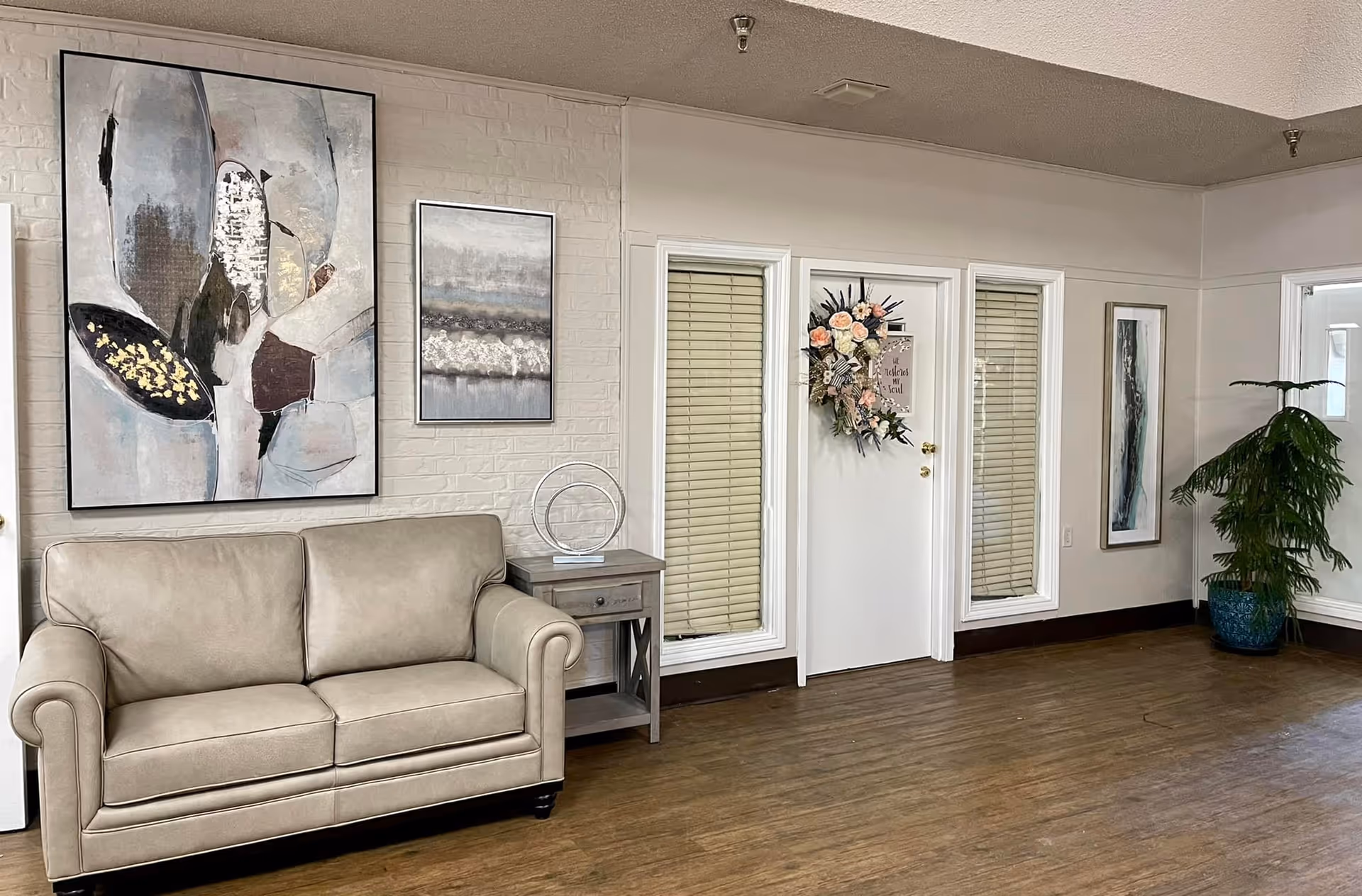 A cozy interior space featuring a beige leather loveseat, a small wooden side table with a modern circular sculpture, and three abstract paintings on the walls. The room has wooden flooring, white walls with a brick texture on one side, and a white door decorated with a floral wreath. There is also a tall green potted plant in a blue pot near the corner.