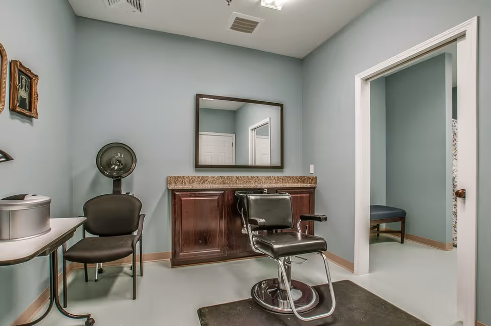 Interior of a small salon or grooming room with a black salon chair on a black mat in front of a counter with a large mirror above it. To the left, there is a black chair with a hair dryer hood behind it and a small table with a silver appliance on top. The walls are painted light blue and there is an open doorway leading to another room with a bench.