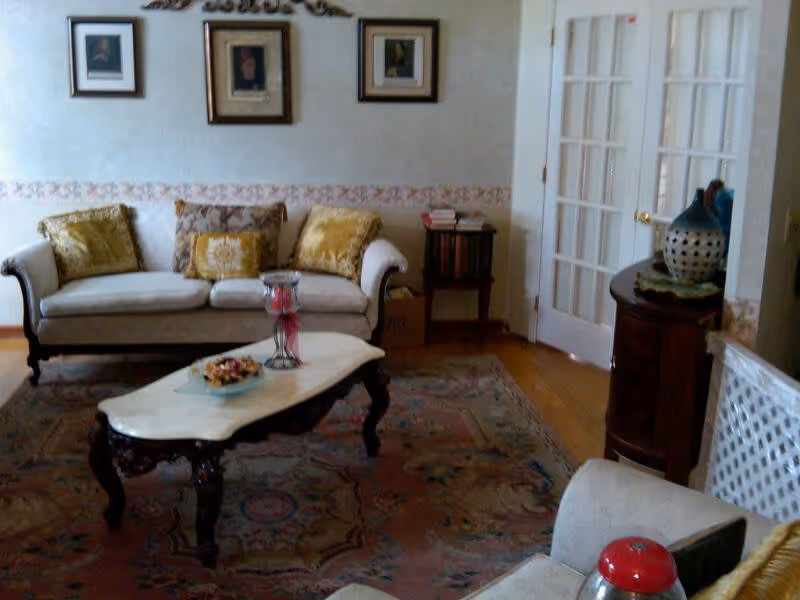 A cozy living room with a white sofa adorned with decorative pillows, a wooden coffee table with a marble top holding a decorative bowl and candle holder, a patterned area rug, framed pictures on the wall, a small side table with books, and a cabinet with a decorative vase near white French doors.