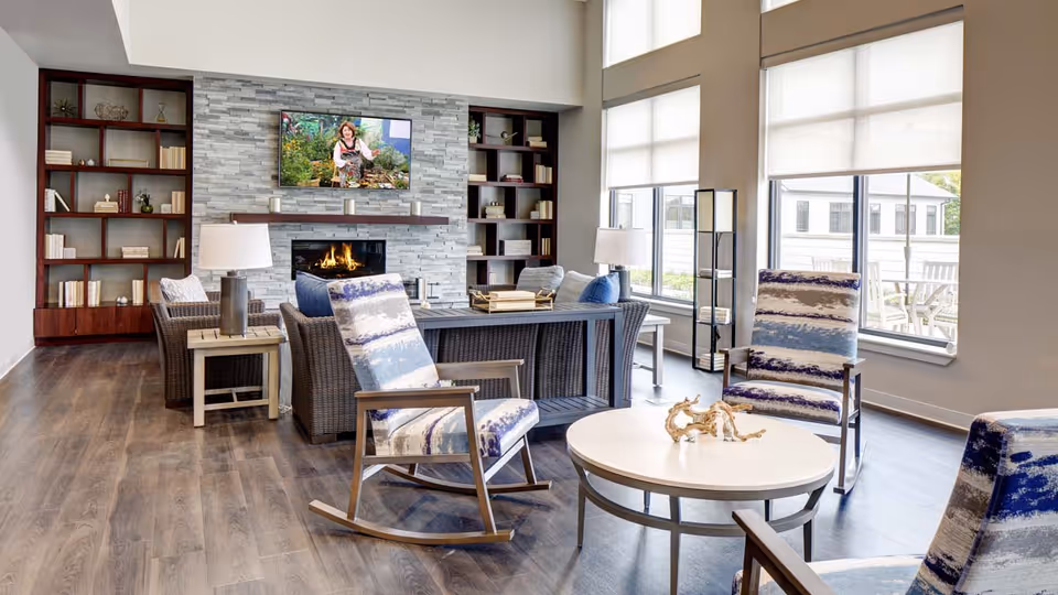 A bright and spacious living room area with large windows letting in natural light. The room features a modern fireplace with a TV mounted above it, flanked by two tall bookshelves filled with books and decorative items. There are comfortable wicker chairs with cushions, two patterned rocking chairs, and a round white coffee table with a decorative piece on it. The floor is wood, and the walls are painted in neutral tones.