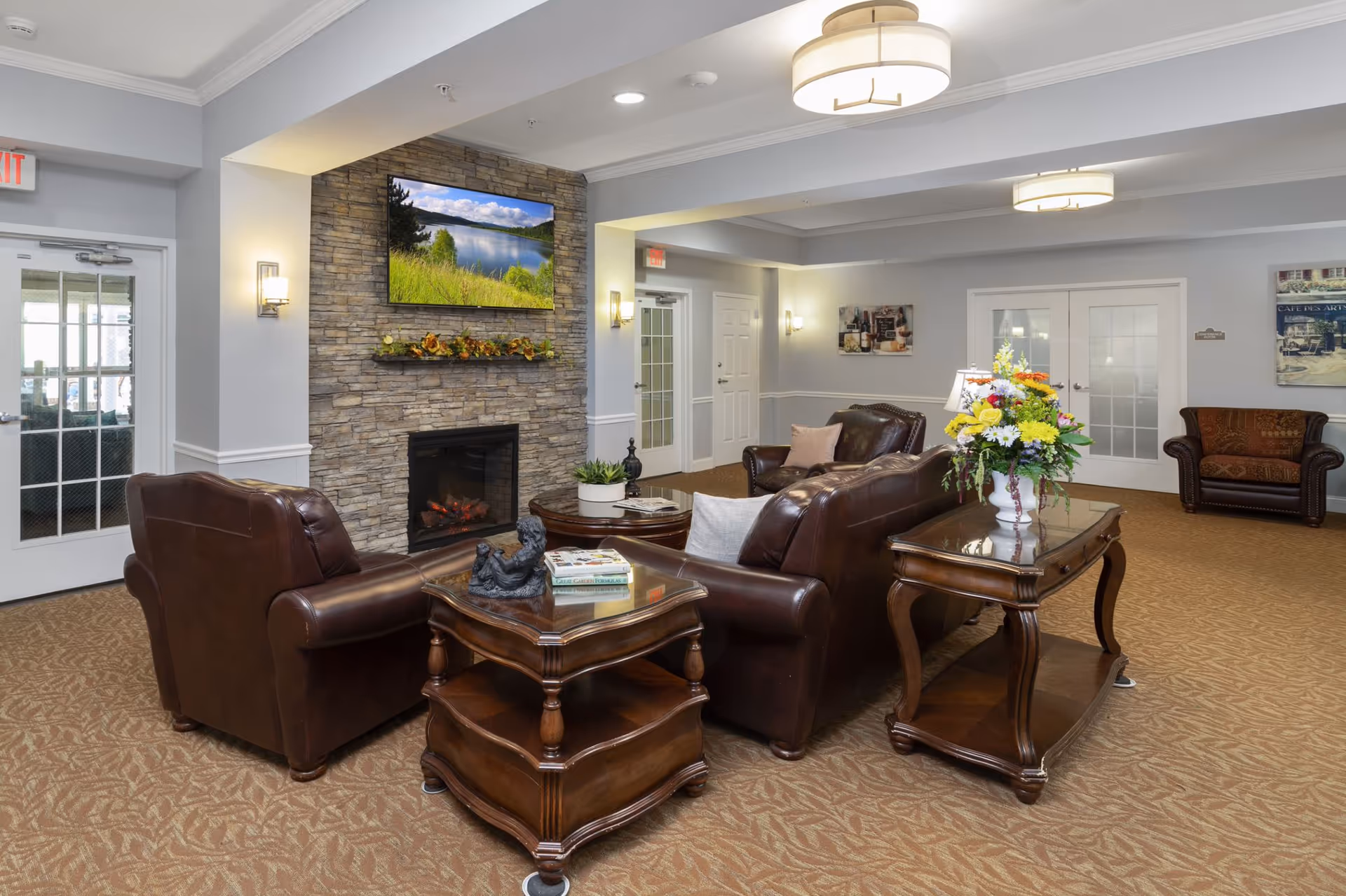 Lounge area with brown leather sofas and chairs arranged around a stone fireplace and wall-mounted TV, wooden side tables and a vase of flowers.