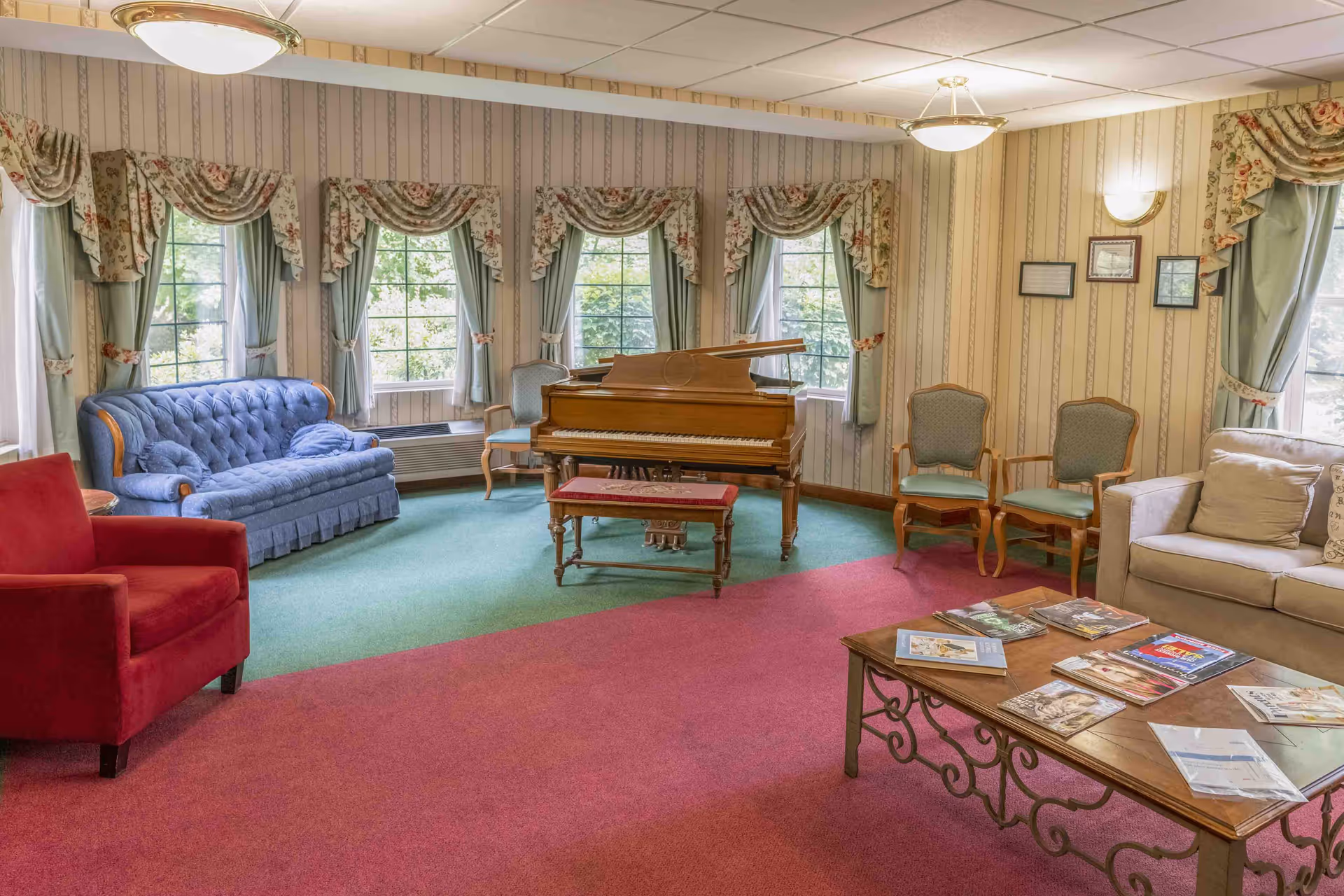 Cozy common lounge with a grand piano, upholstered sofas and chairs around a coffee table in front of multiple windows.
