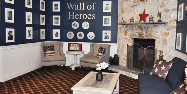 A cozy living room area with a stone fireplace decorated with a red star and small trophies on the mantel. There are two beige armchairs with American flag pillows around a small white table with a 'Heroes Welcome' sign. A dark blue couch with star-patterned pillows faces a white coffee table with a small flower arrangement. The walls feature a 'Wall of Heroes' display with framed black and white photos and decorative plates.