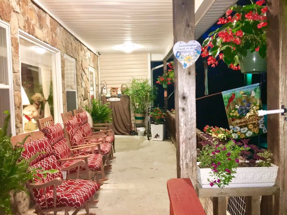 A covered outdoor porch area at night with several wooden rocking chairs featuring red patterned cushions lined up against a stone wall with windows. The porch is decorated with various potted plants and hanging flower baskets. A heart-shaped welcome sign hangs on a wooden post, and a colorful garden flag is visible. The area is well-lit with ceiling lights.