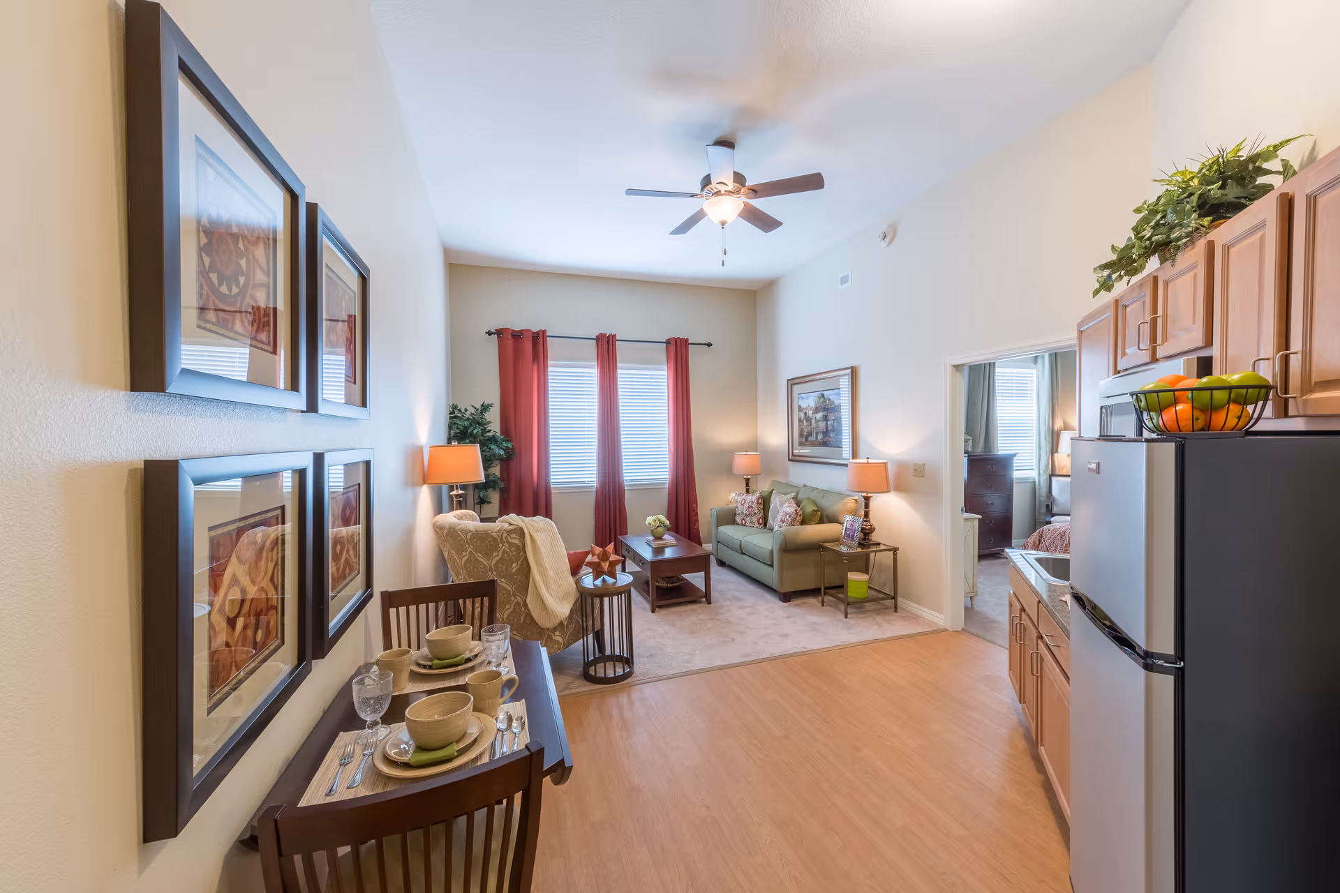 Open-plan living area with a small dining table, kitchenette on the right, and a seating area with a green sofa and red curtains.