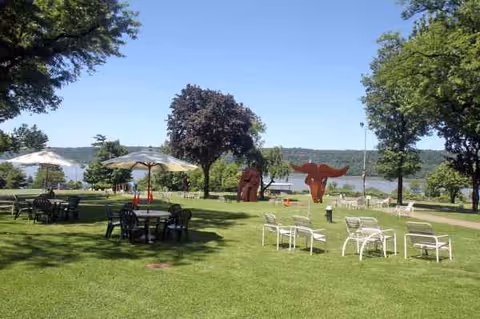 Sunny lakeside lawn with umbrellas, seating, tables, and red sculptures.