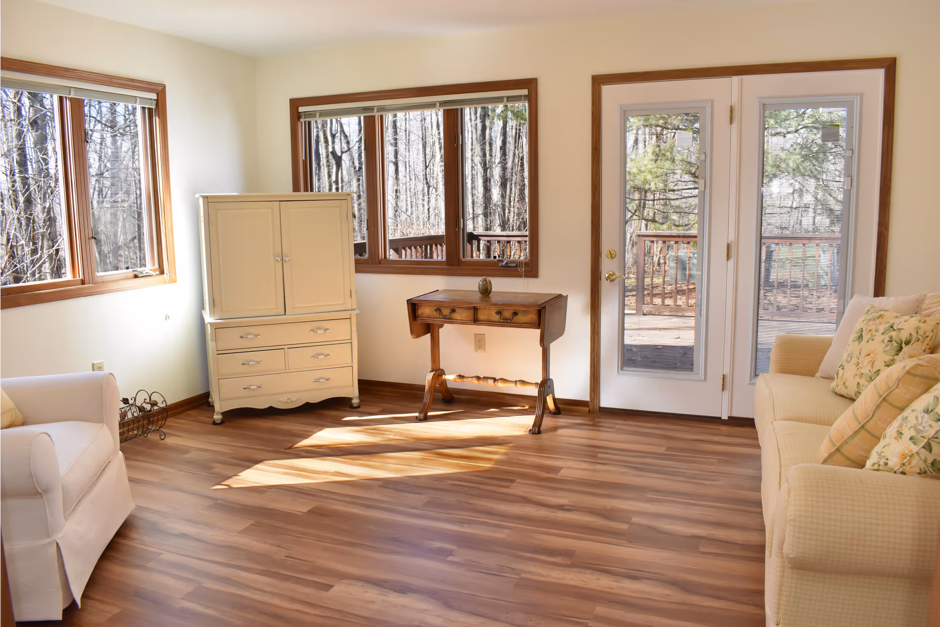 A bright living room with wooden flooring, two large windows, and a glass door leading to an outdoor deck. The room contains a cream-colored armchair on the left, a cream-colored sofa with floral and plaid cushions on the right, a small wooden table in the center, and a cream-colored cabinet near the windows. Sunlight streams through the windows, illuminating the room.