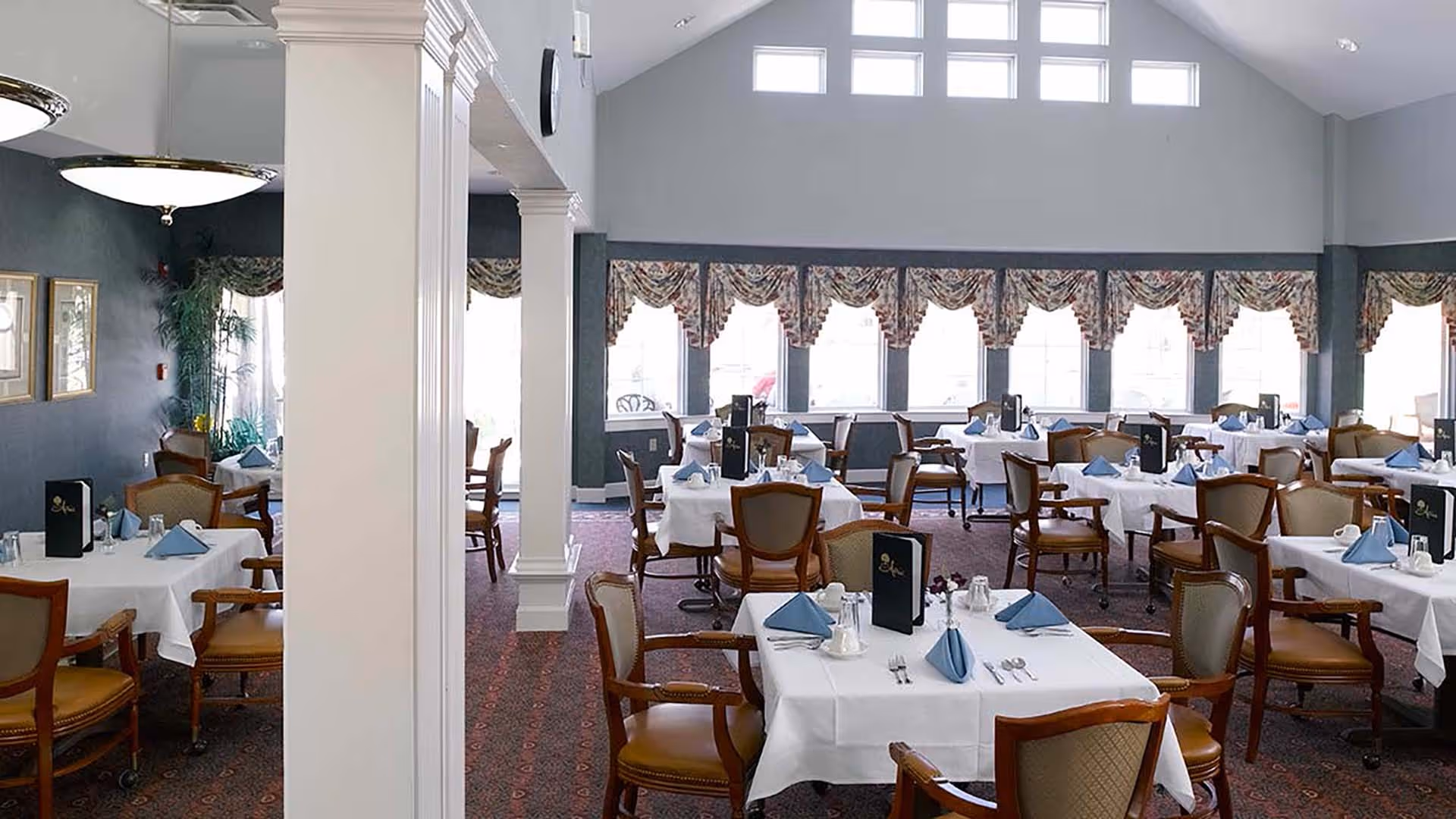 A spacious dining room with multiple tables covered in white tablecloths, each set with blue folded napkins, silverware, salt and pepper shakers, and black menus. The room has large windows with patterned valances, high ceilings with small square windows near the top, and wooden chairs with cushioned seats and backs.
