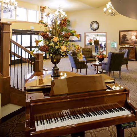 Lobby of a senior living community featuring a polished grand piano with a floral arrangement, seating area, and chandeliers.