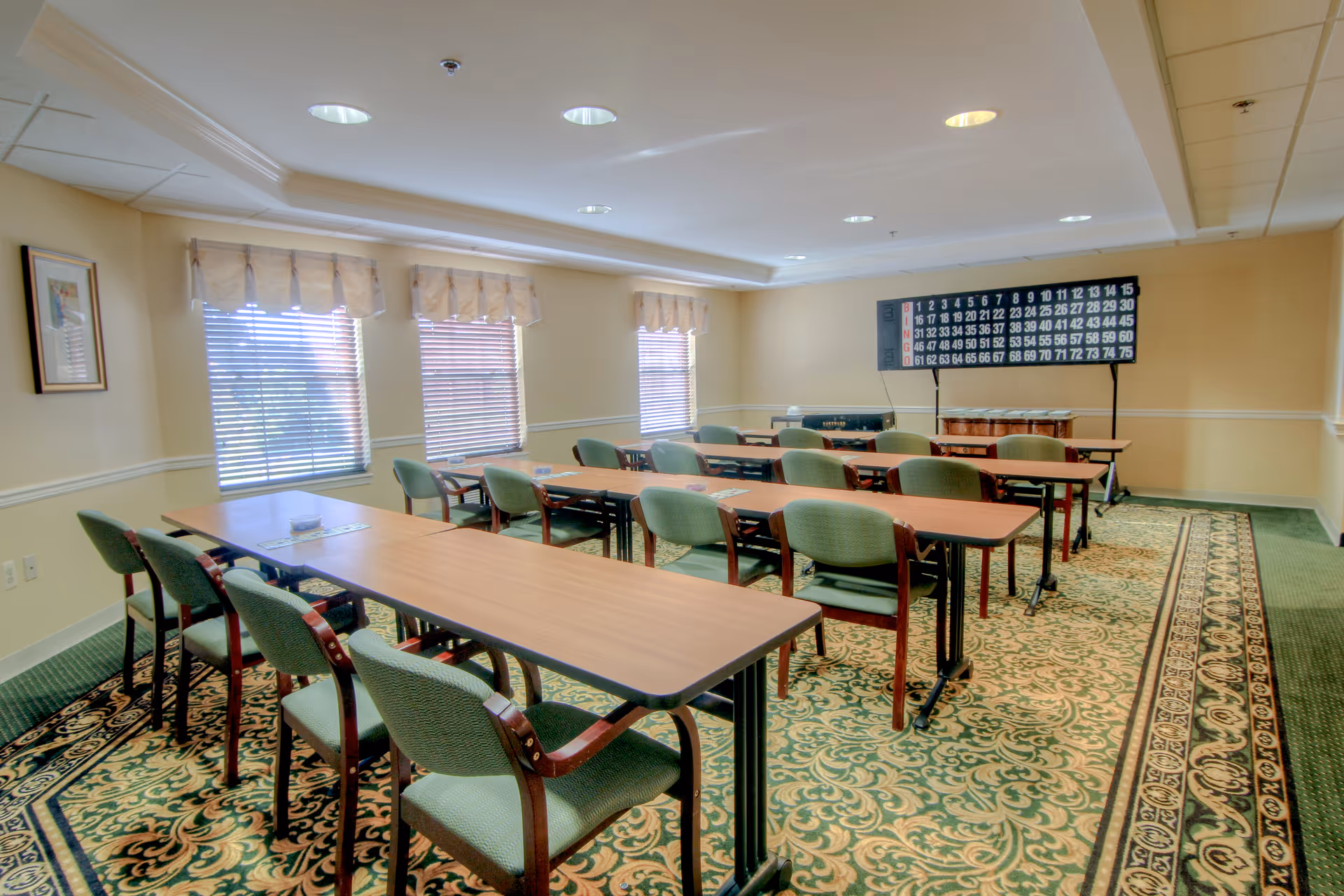 A well-lit room with several rectangular tables arranged in rows, each accompanied by green cushioned chairs. The room has three windows with blinds and valances, a patterned carpet, and a large bingo board on a stand at the far end.