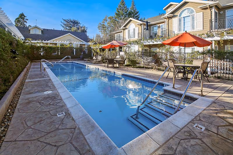 Outdoor rectangular swimming pool with steps, lounge chairs and tables with red umbrellas in a courtyard of a two-story residential building.