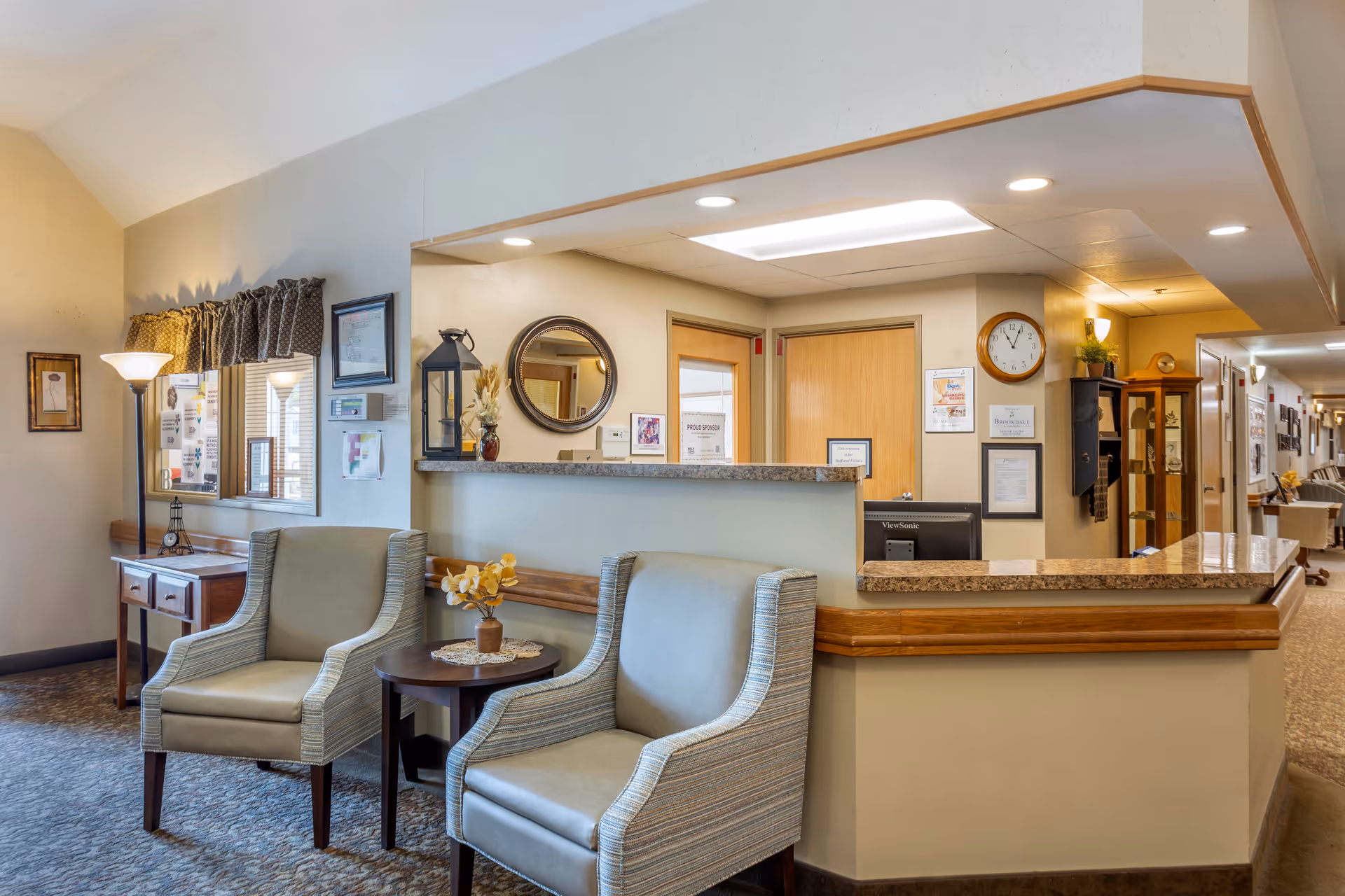 A reception area in a senior living facility featuring a front desk with a granite countertop, two upholstered armchairs with a small round table between them holding a vase with yellow flowers, a floor lamp, a wooden side table with drawers, a window with patterned valance, and a hallway leading to other rooms. The walls are decorated with framed pictures, a round mirror, a clock, and certificates.