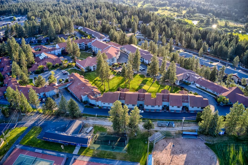 Aerial view of Fairwood Retirement Village & Assisted Living showing multiple buildings with red roofs surrounded by tall trees and green lawns. The facility is set in a wooded area with roads and parking spaces visible, along with tennis courts and a baseball field in the foreground.