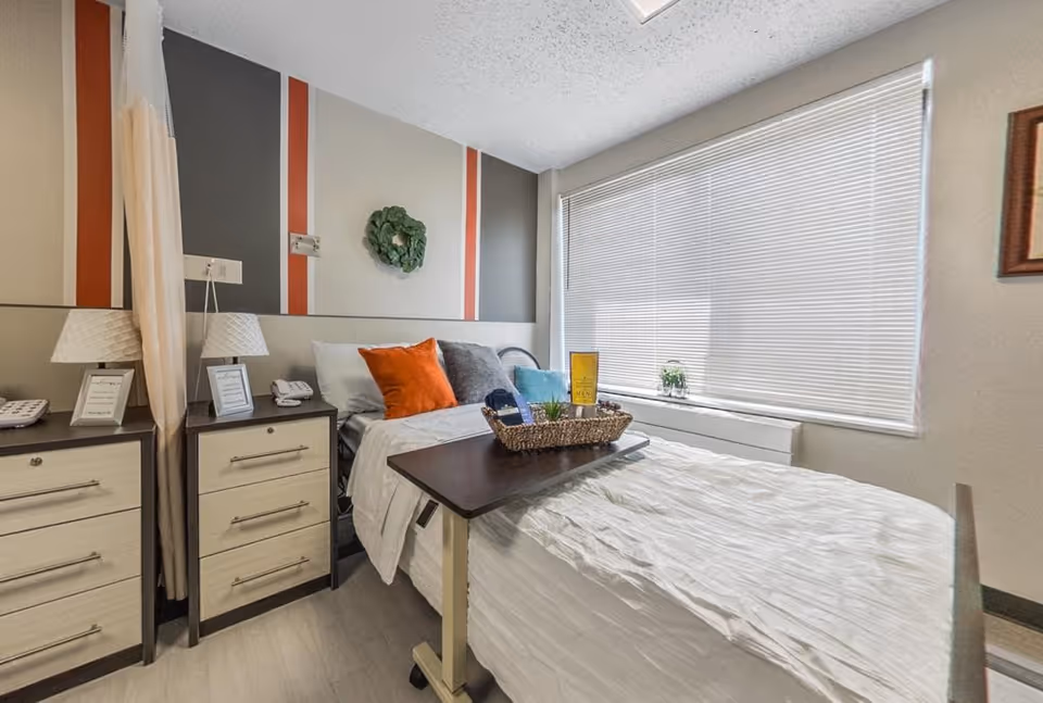A bright and clean bedroom in a rehabilitation and nursing center featuring a single bed with white bedding and colorful pillows, a bedside table with a lamp and phone, a small rolling table with a basket of items on the bed, a large window with blinds, and a decorative wreath on the wall.