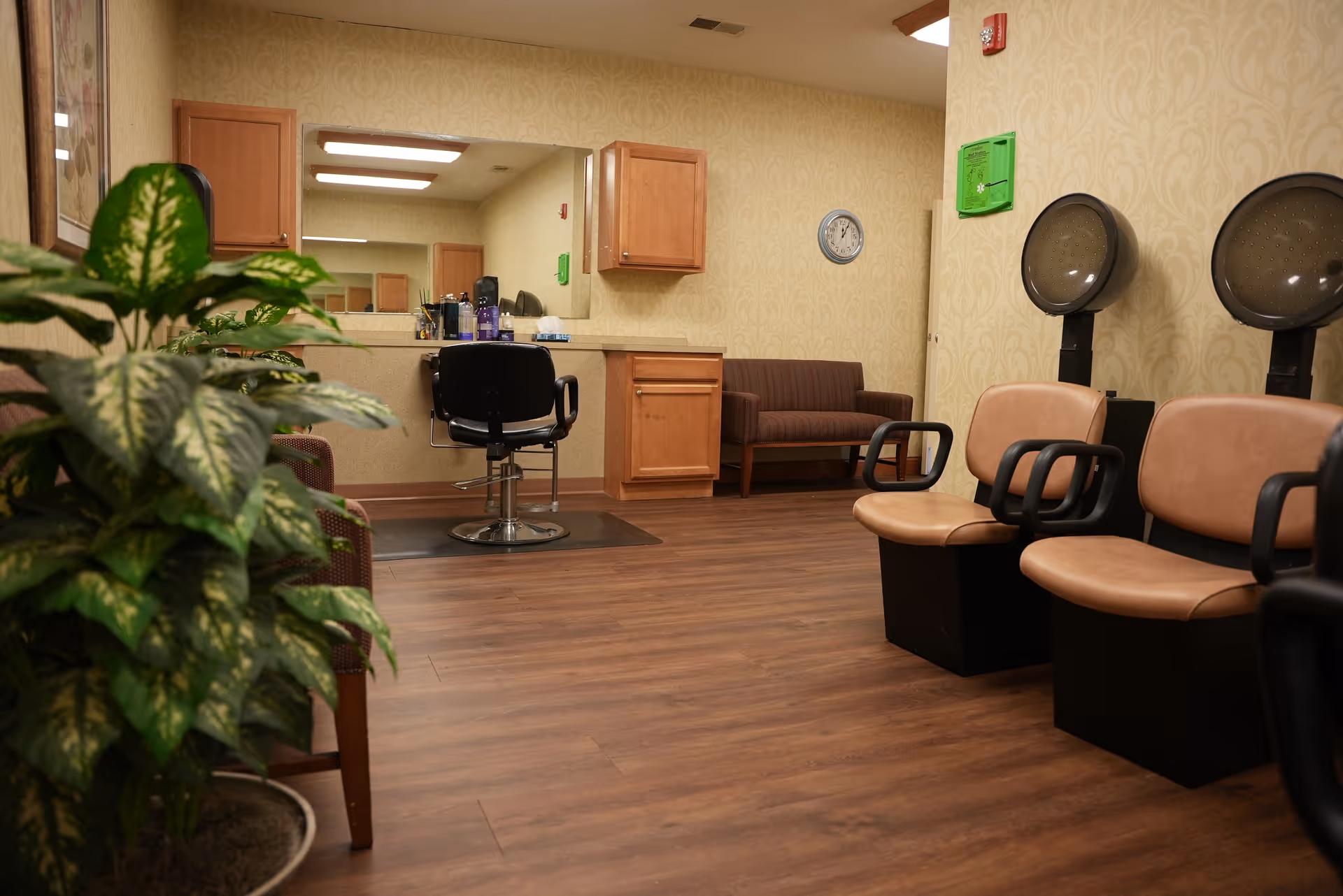 Salon area inside an assisted living facility with a styling chair and mirror, hooded hair dryers, seating, and plants.