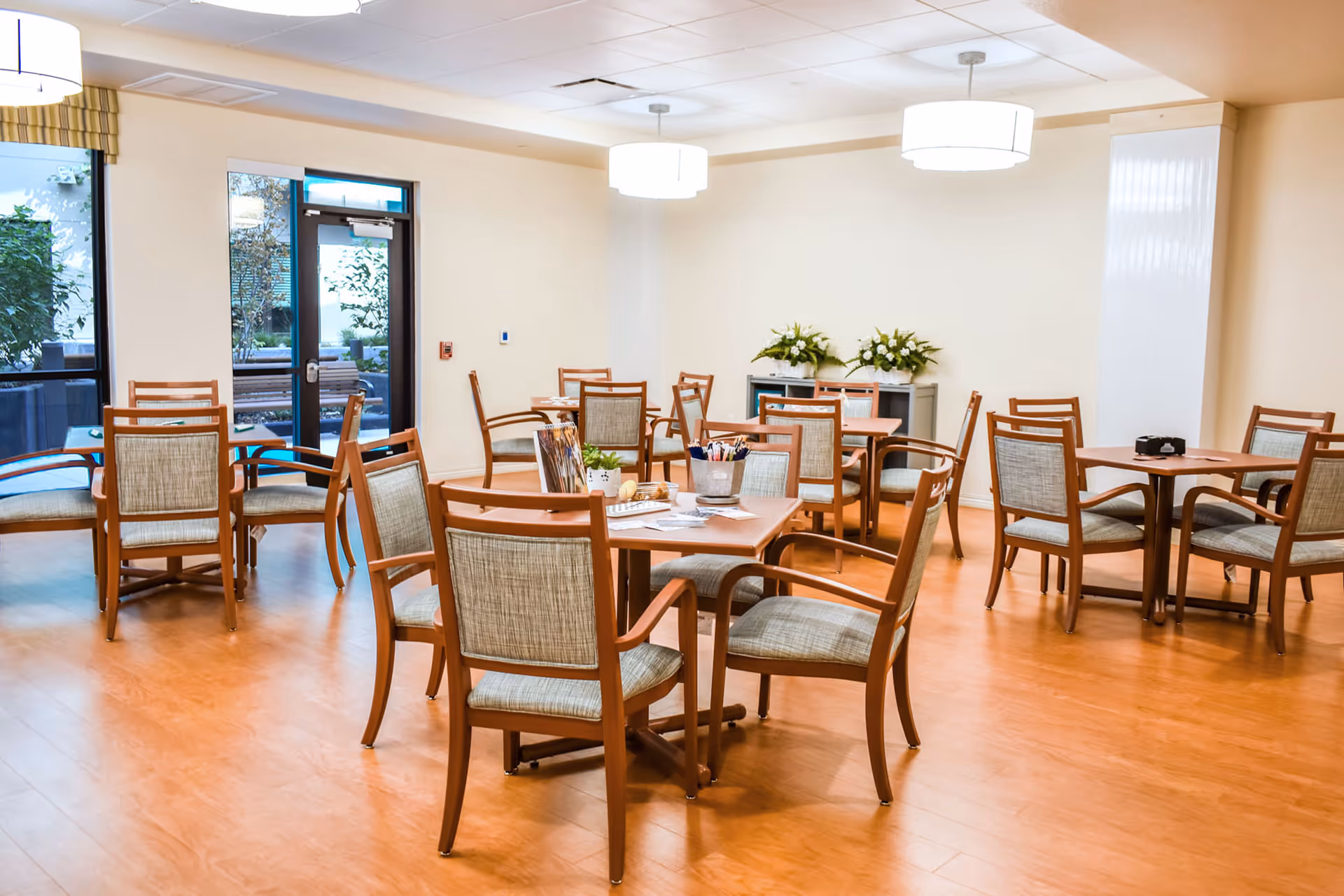A bright and clean dining room with multiple wooden tables and cushioned chairs arranged neatly. The room has large windows and a glass door letting in natural light, with plants visible outside. The floor is wooden, and there are modern ceiling lights and floral arrangements on a side table.