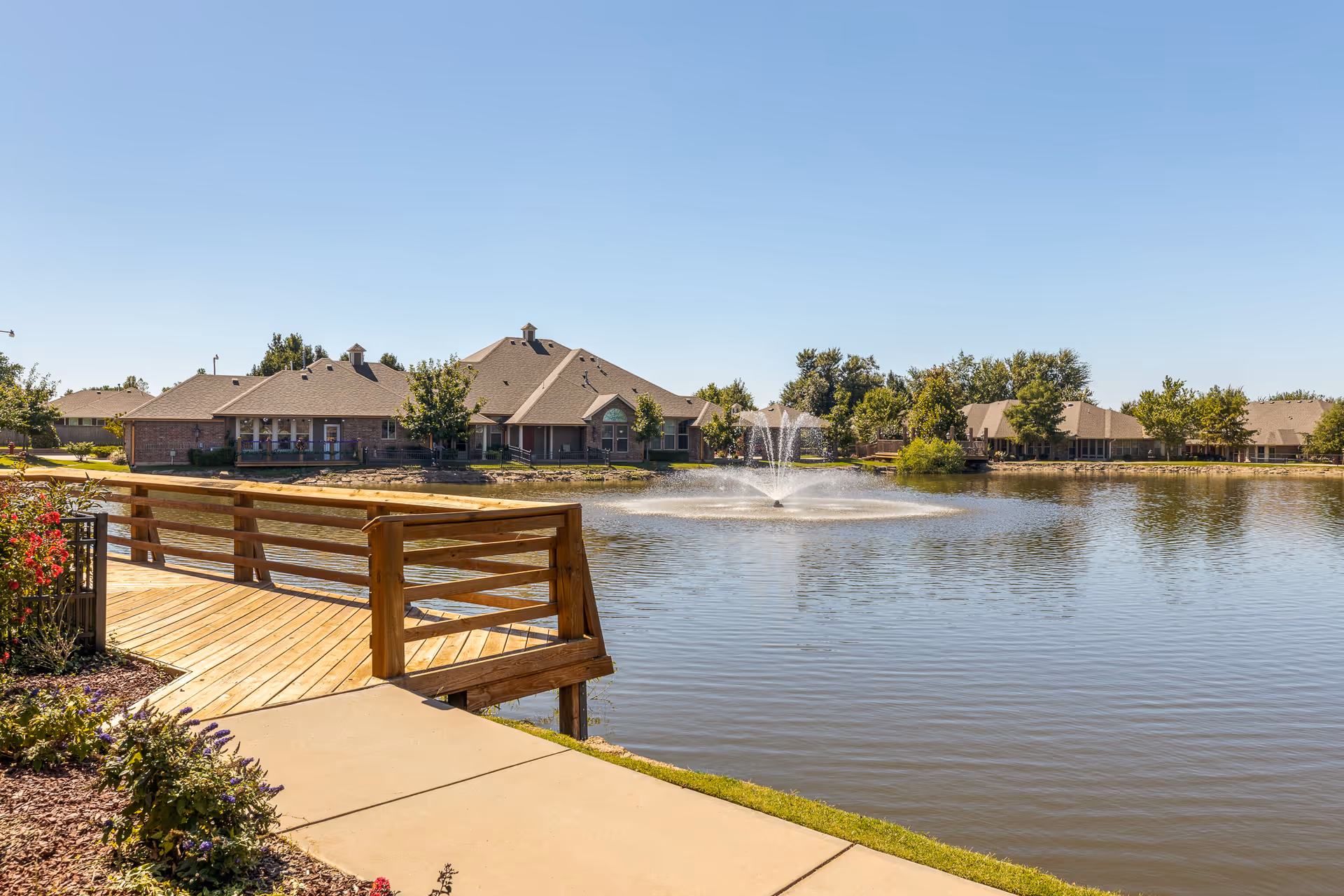 Wooden boardwalk beside a pond with a central fountain and single-story brick buildings in the background under a clear blue sky.