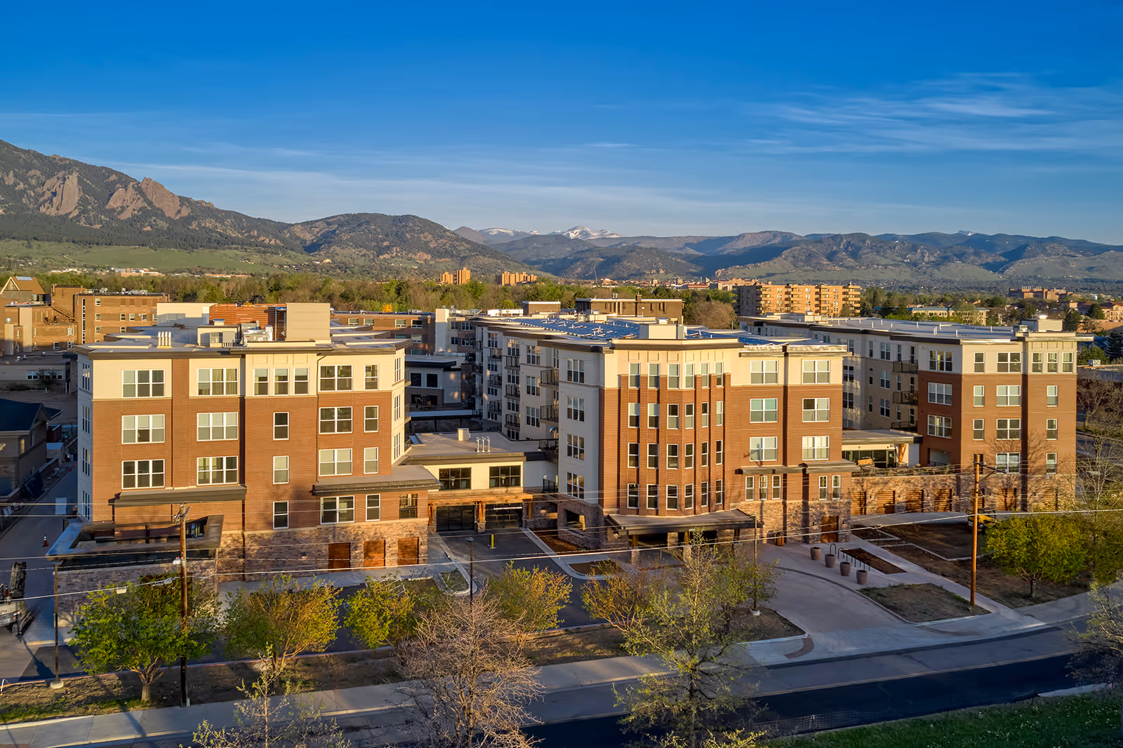 Aerial view of Frasier Retirement Community, a multi-story residential building complex with a mix of brick and light-colored exterior walls, set against a backdrop of mountains under a clear blue sky.