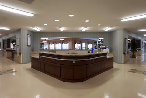Central reception desk in a senior living facility lobby with wood paneling, overhead lights, and seating visible in the background.