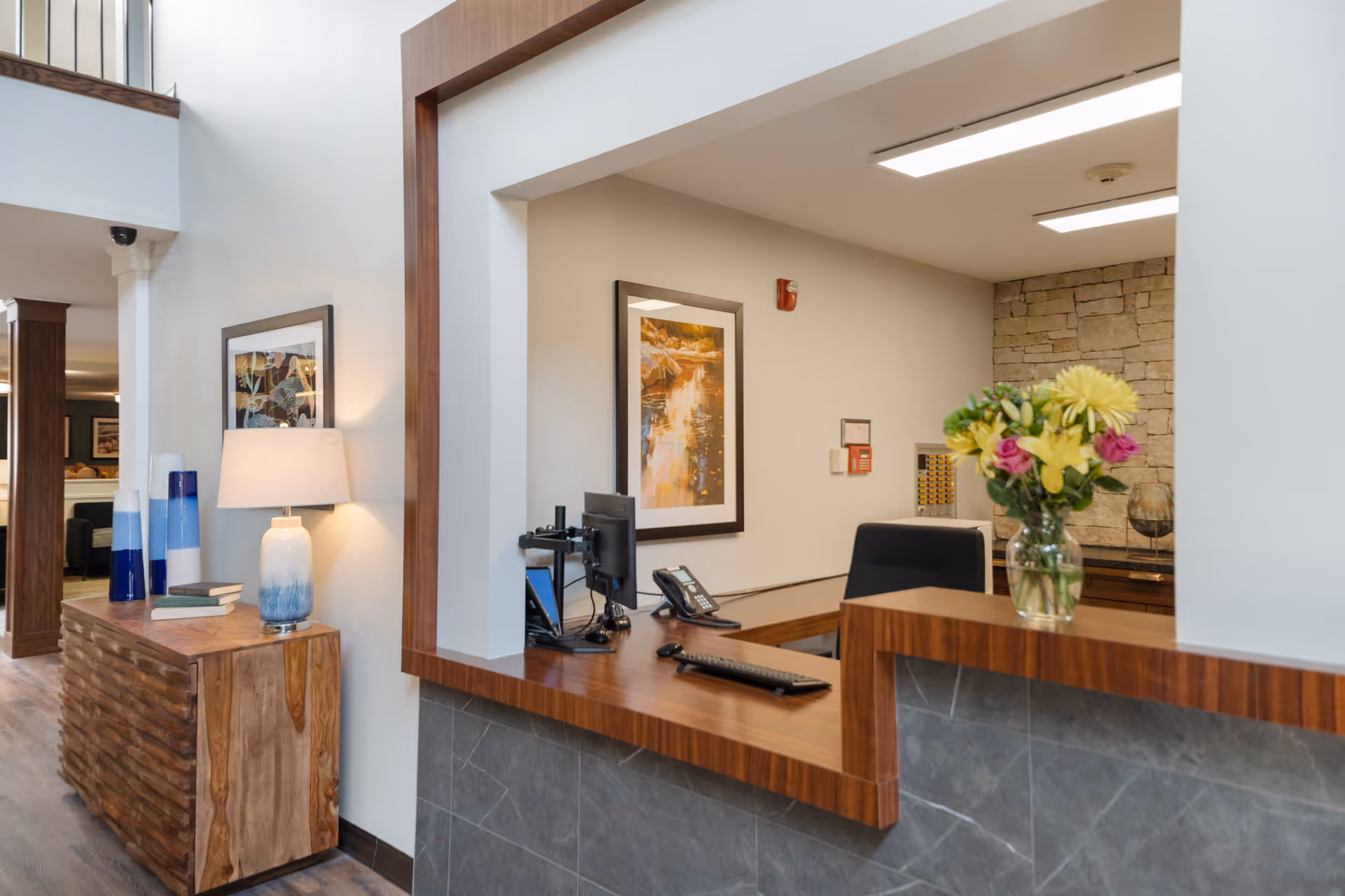 Reception desk area in a senior living facility with a wooden counter, a computer, a phone, and a vase of flowers. Behind the desk is a framed picture on the wall and a stone accent wall. To the left, there is a wooden side table with a lamp, books, and decorative vases. The space has a warm and welcoming atmosphere.