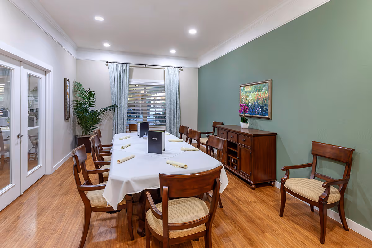 A formal dining room with a long table set with napkins, wooden chairs, a green accent wall, and a sideboard with artwork and a plant.