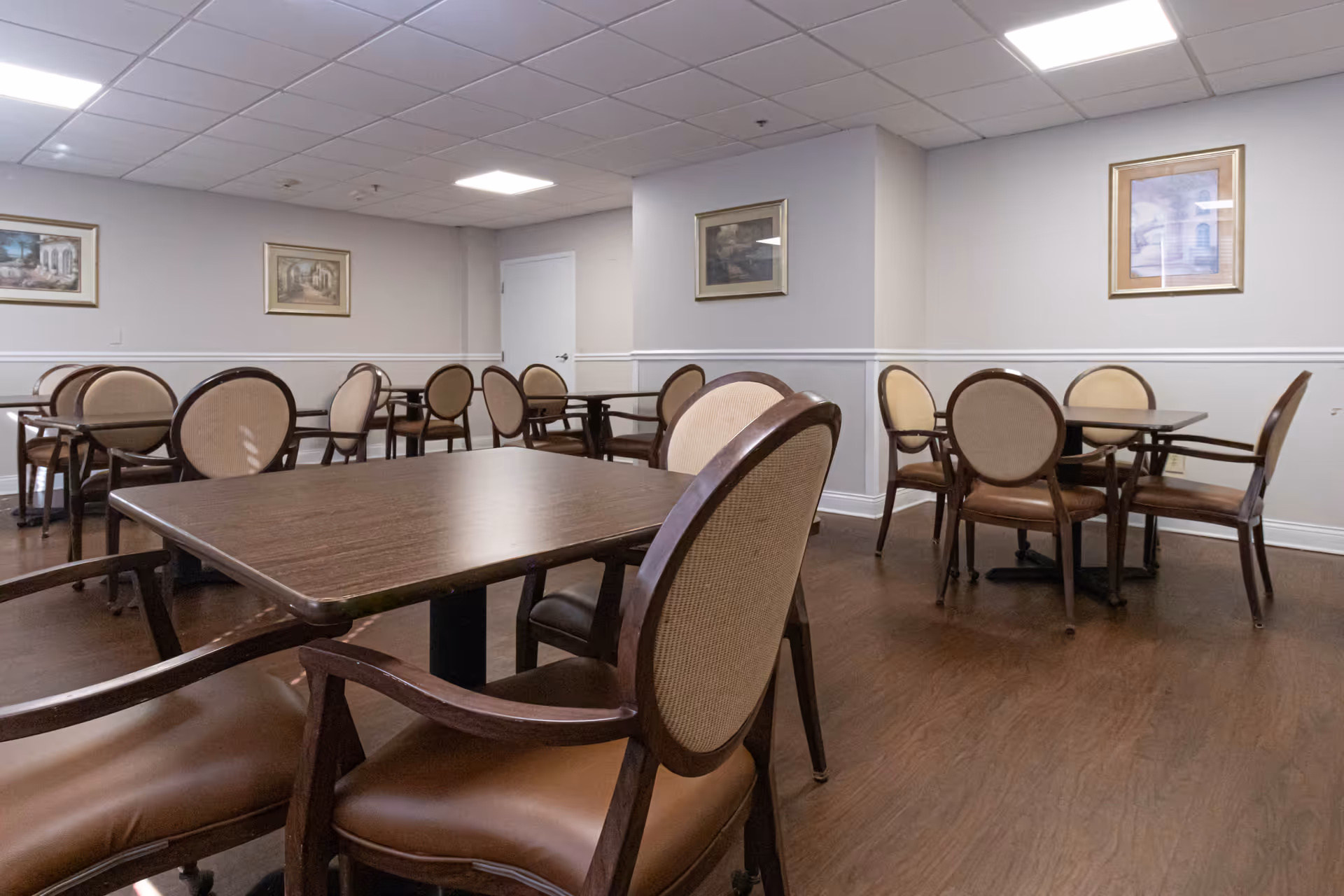 A dining room with multiple wooden tables and cushioned chairs arranged neatly. The room has light-colored walls adorned with framed artwork and a white door in the background. The floor is wooden, and the ceiling has recessed lighting panels.