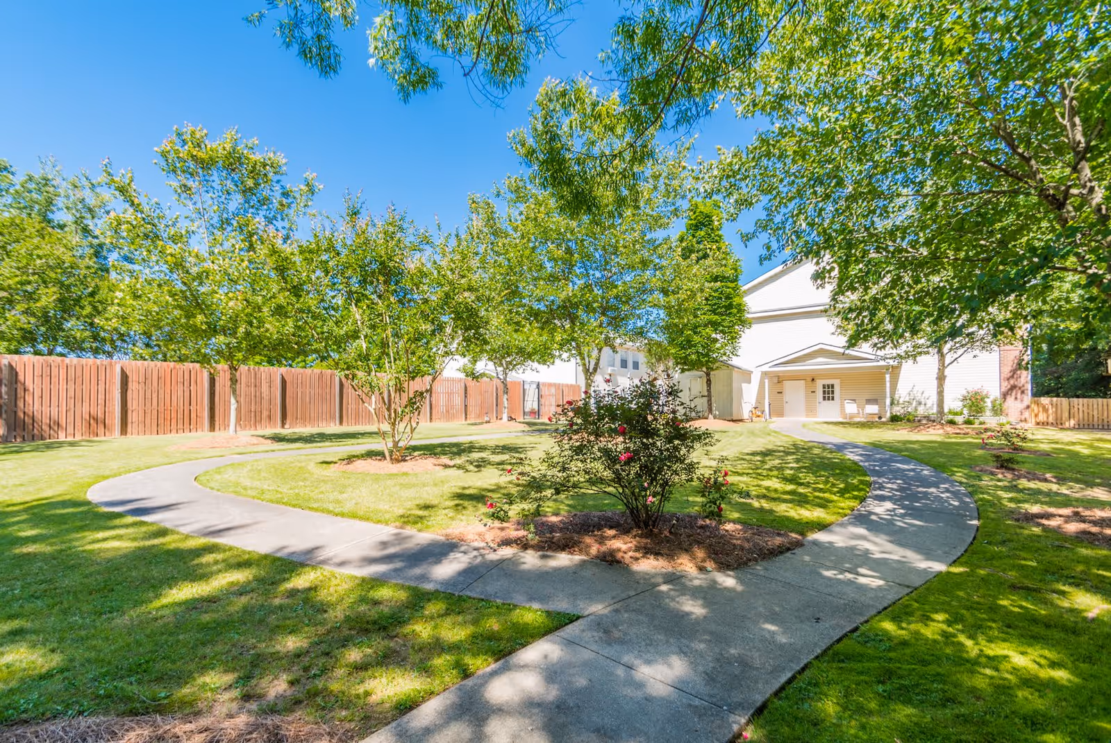 A sunny outdoor garden area with a curved concrete pathway surrounded by green grass, trees, and flowering bushes. A white building with a covered porch and two chairs is visible in the background, along with a wooden fence enclosing the space.