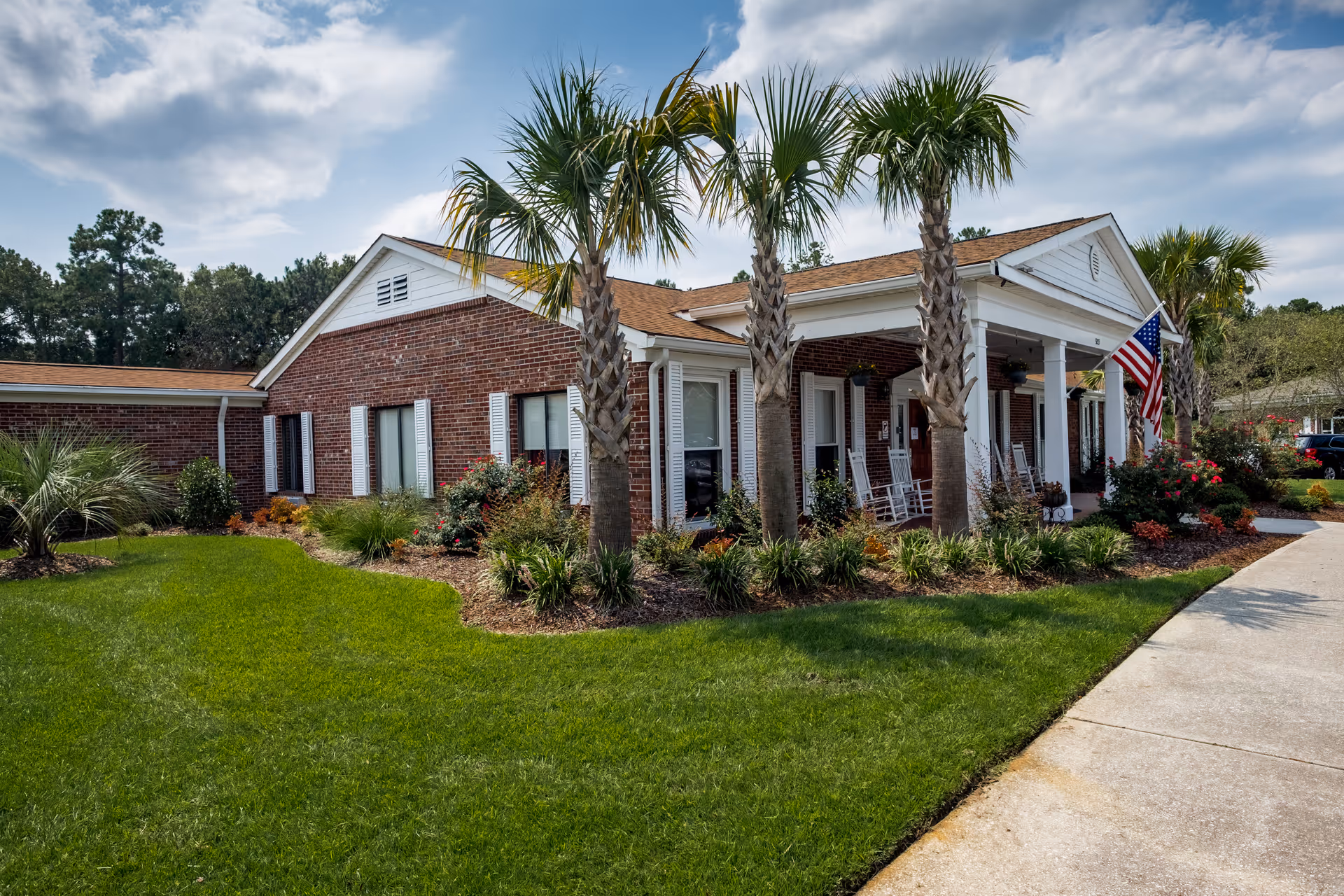 Exterior view of a single-story brick building with white trim and a covered porch featuring rocking chairs. The building is surrounded by well-maintained landscaping including palm trees, shrubs, and green grass under a partly cloudy sky. An American flag is displayed near the porch.