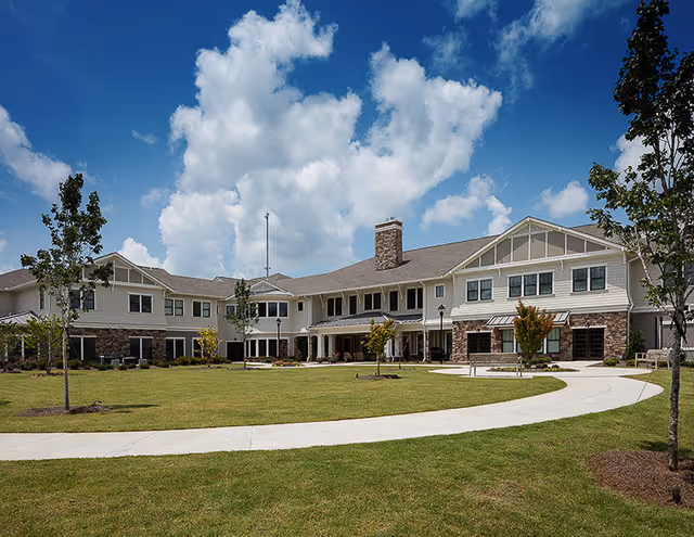 Exterior view of a large two-story senior living facility building with a stone chimney, multiple windows, and a covered entrance. The building is surrounded by a well-maintained lawn with young trees and a curved concrete walkway under a partly cloudy blue sky.