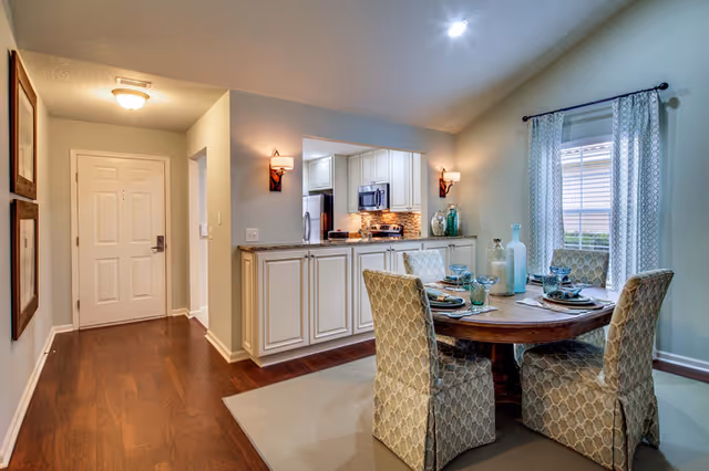 A bright interior dining area with a round table and four upholstered chairs adjacent to a pass-through kitchen and entry hallway.