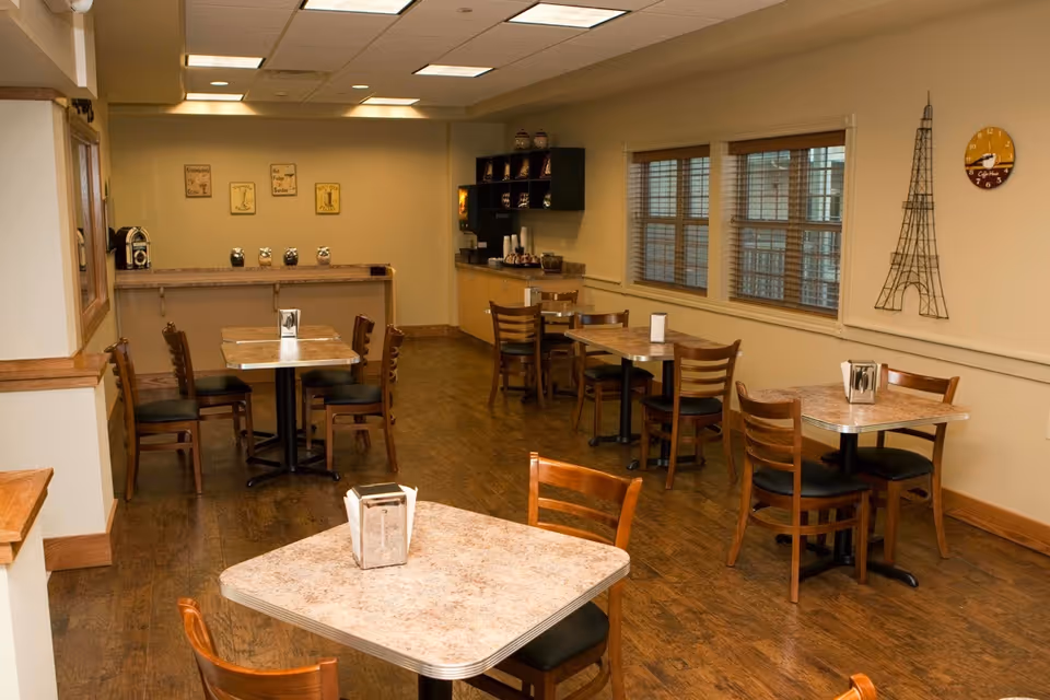 A small dining area with several square tables and wooden chairs arranged neatly. The room has wooden flooring, beige walls, and windows with blinds. On one wall, there is a decorative wire sculpture resembling the Eiffel Tower and a round clock. In the back corner, there is a counter with coffee and condiment supplies.