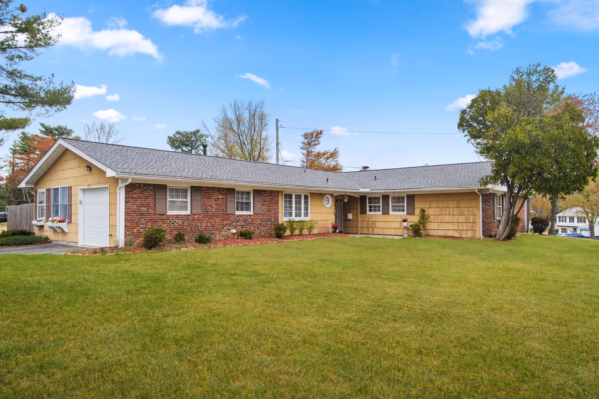 Single-story assisted living facility building with a combination of brick and yellow siding exterior, a gray shingled roof, several windows with brown shutters, a garage door on the left side, and a well-maintained green lawn in front under a blue sky with some clouds.