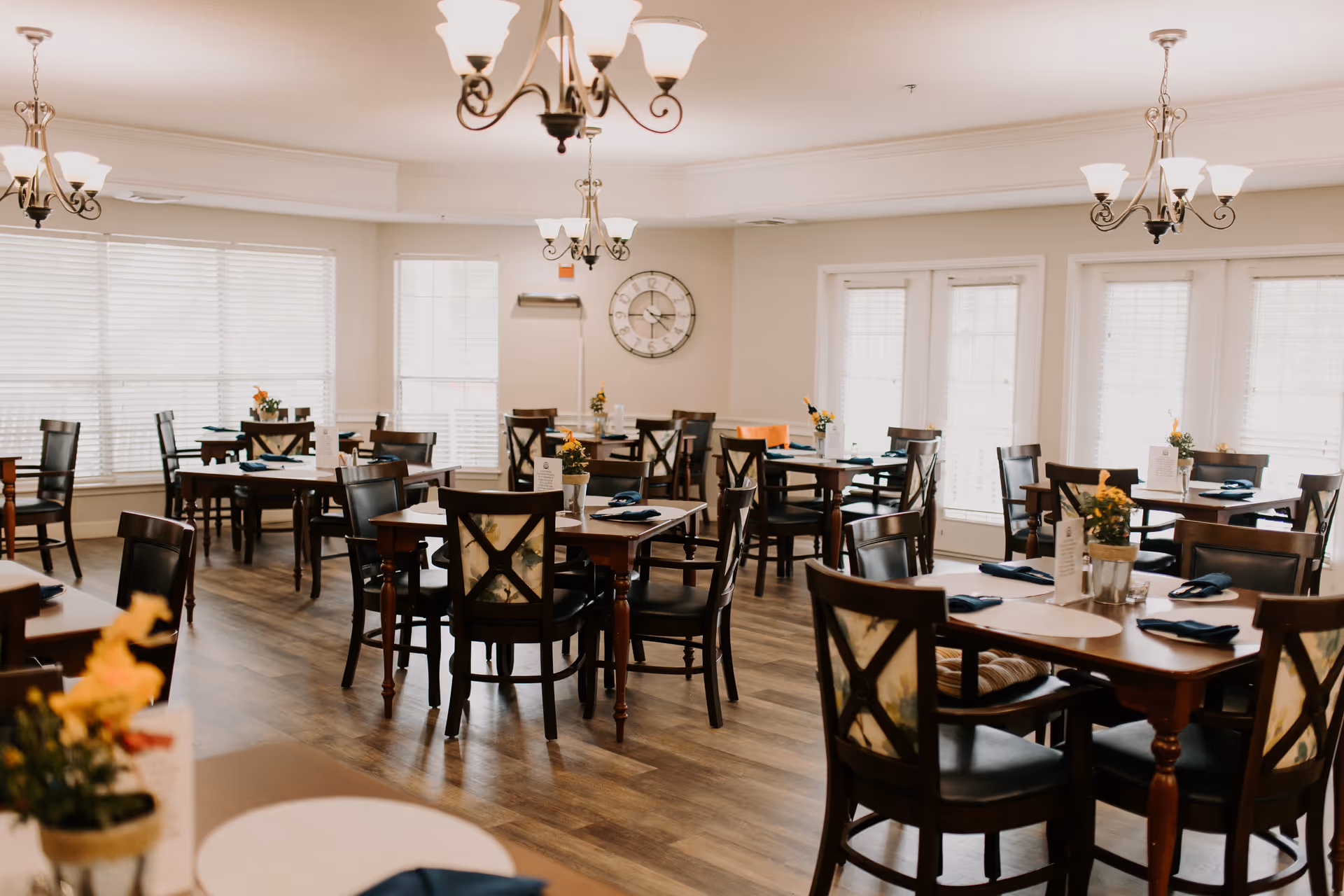 Well-lit dining room with multiple wooden tables and chairs set with napkins and small floral centerpieces under chandeliers.