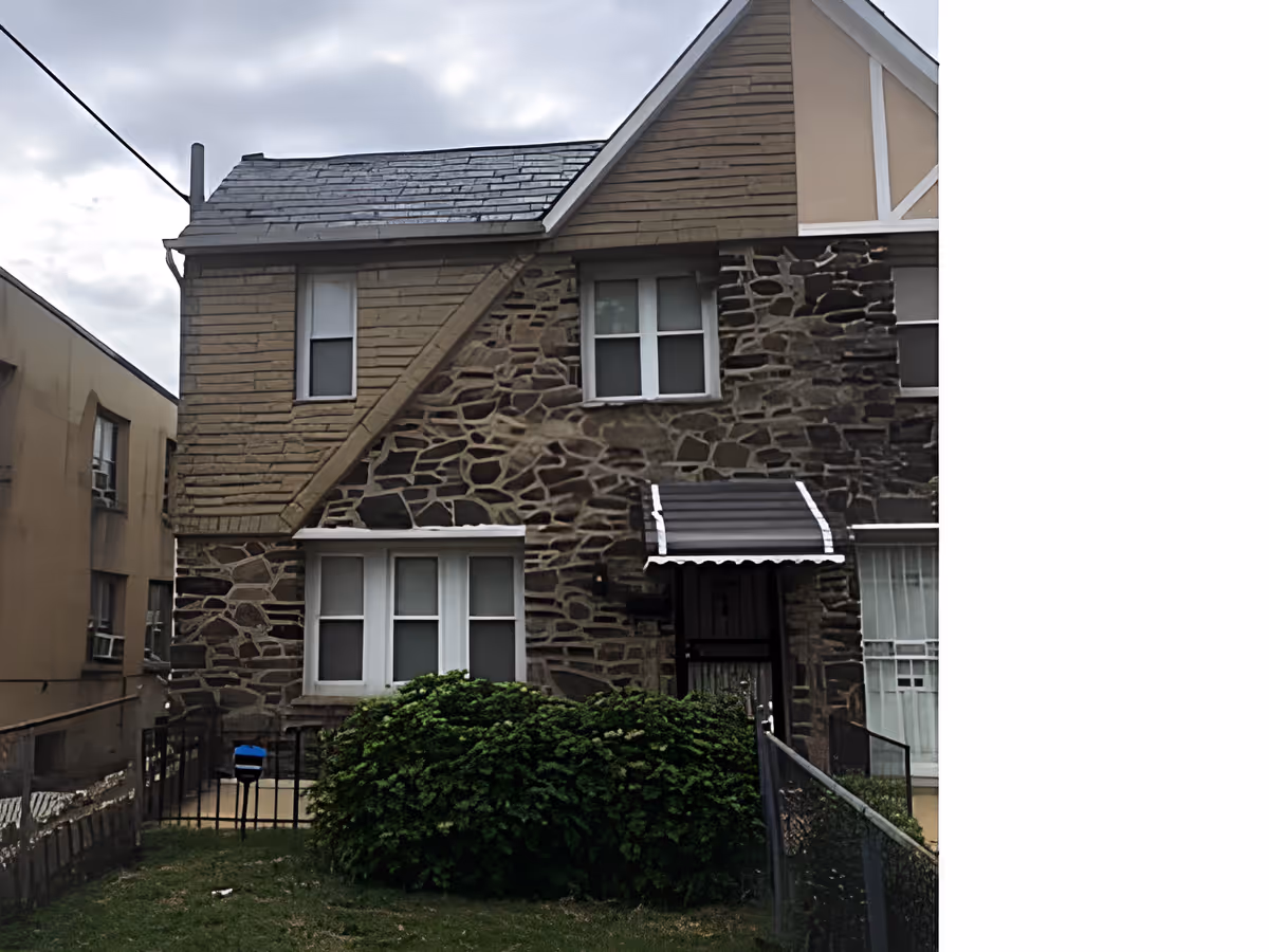 Exterior view of a two-story stone and wood house with a small front yard, green bushes, and a black metal fence. The house has multiple windows and a small awning above the front door. The sky is cloudy.