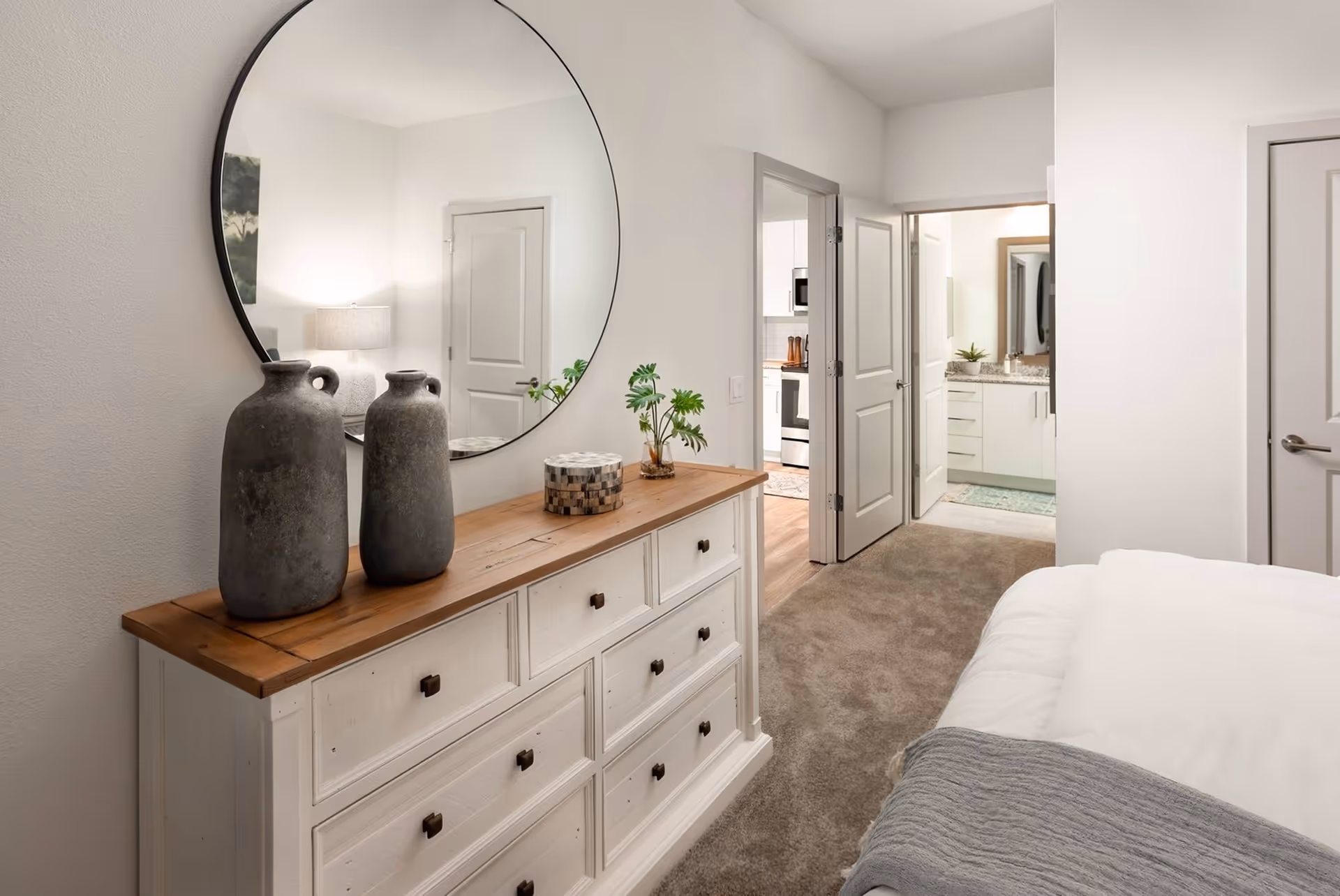 Bedroom interior with a white wooden dresser topped by two gray vases and a round mirror, a bed at right, and open doors leading to a bathroom and kitchen.