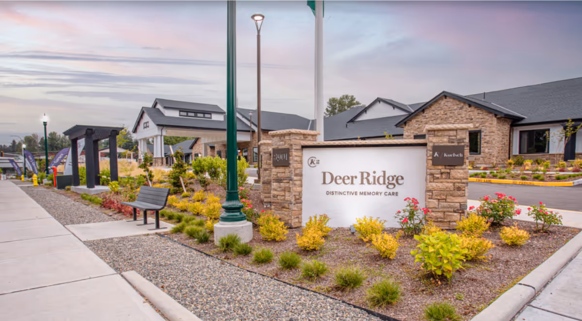 Exterior view of Deer Ridge Distinctive Memory Care facility with a stone and white sign, landscaped garden with bushes and flowers, a bench, and a covered entrance to the building under a partly cloudy sky.