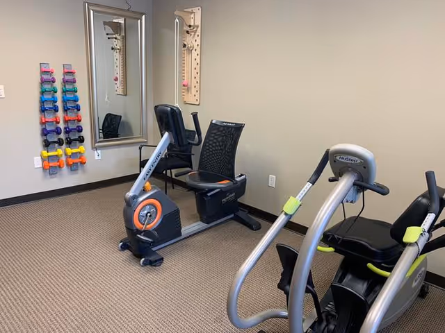 A small exercise room with two stationary recumbent bikes, a rack of colorful dumbbells mounted on the wall, two large mirrors, and a pegboard with pegs for exercises. The room has beige walls and carpeted flooring.