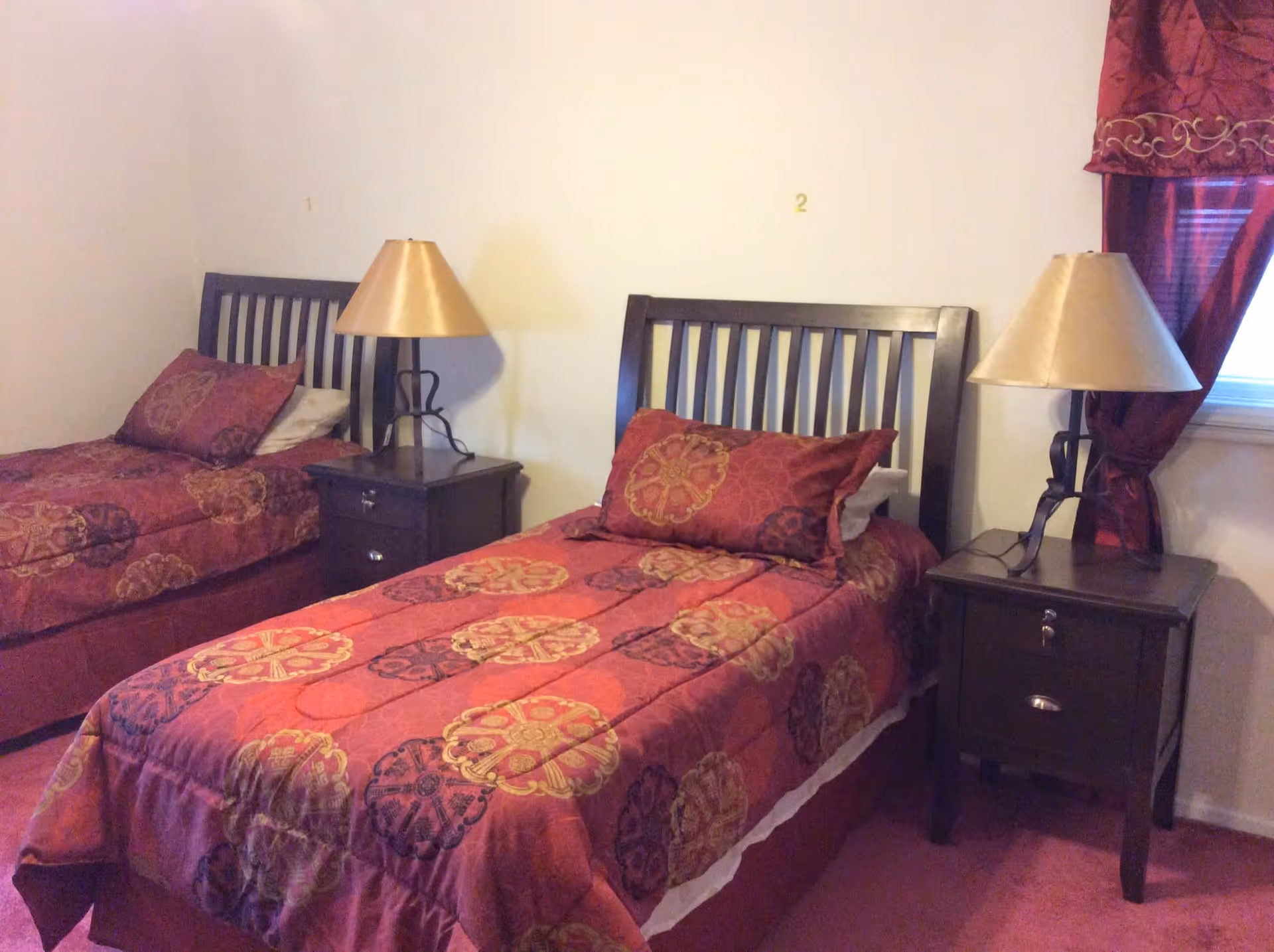 A bedroom with two single beds featuring dark wooden headboards and red patterned bedspreads. Each bed has a matching pillow and a nightstand with a lamp. There is a window with red curtains on the right side of the room.