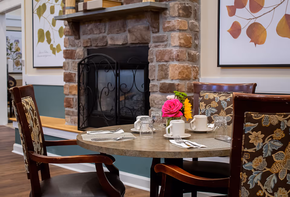 A round dining table set for four with placemats, utensils, mugs, and glasses in front of a stone fireplace with a black metal screen. The chairs have floral patterned cushions, and there are framed botanical prints on the walls.