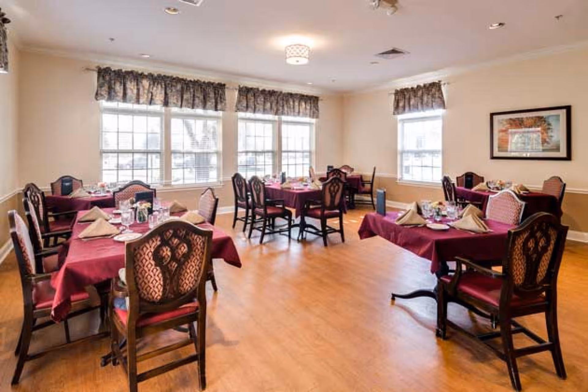 A dining room with several tables covered in burgundy tablecloths, each set with beige folded napkins, plates, glasses, and silverware. The room has large windows with floral valances, wooden chairs with patterned upholstery, light-colored walls, and a wooden floor. A framed picture hangs on one wall.