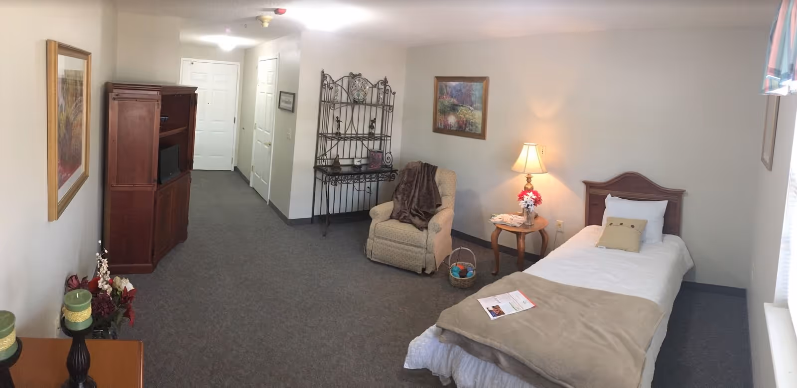 A senior living facility bedroom with a single bed covered in white and beige bedding, a wooden headboard, and a small pillow. Next to the bed is a wooden side table with a lamp and a flower arrangement. There is a comfortable armchair with a blanket draped over it, a decorative metal shelving unit, and a wooden cabinet with a TV. The room has carpeted flooring, framed artwork on the walls, and a window with a valance letting in natural light.