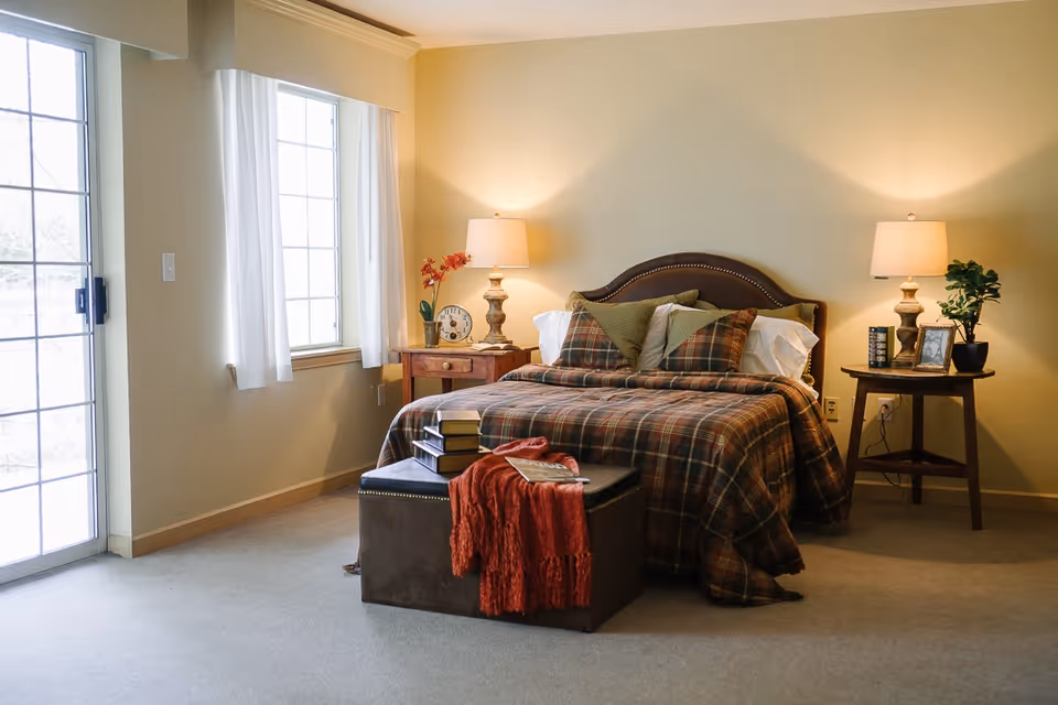 Sunlit bedroom with a plaid-covered bed, bedside tables with lamps, a window, and a sliding glass door.