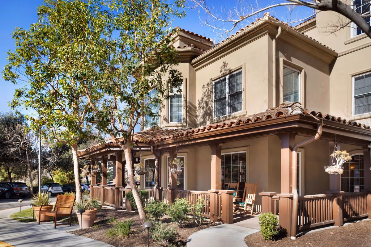 Front exterior of a two-story senior living building with a covered porch, rocking chairs, trees, and potted plants.