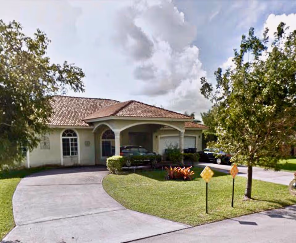 Front exterior view of a single-story building with a tiled roof, arched windows, and a driveway leading to a covered entrance. There are trees and shrubs around the building, and a car is parked under the covered area.