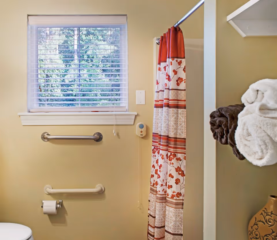 A bathroom interior with a window and blinds, two grab bars, a floral shower curtain, toilet paper, and folded towels on a shelf.
