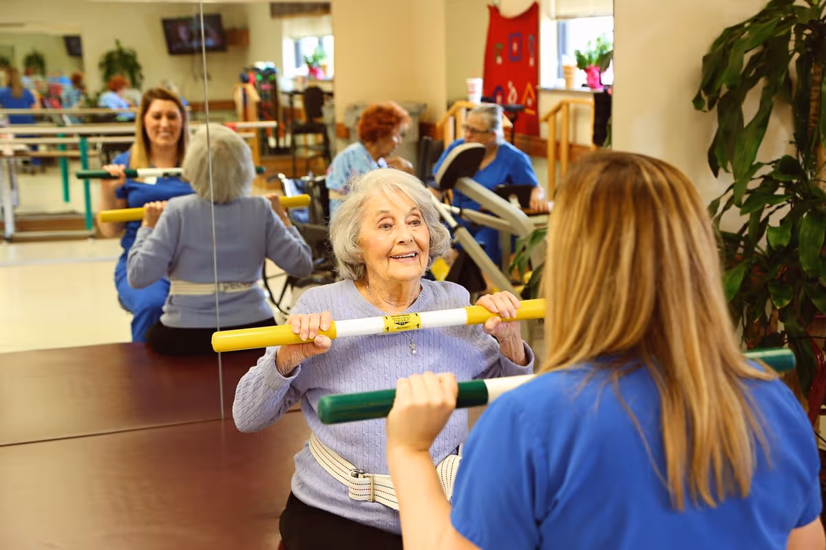 An elderly woman in a light purple sweater is exercising with a yellow and white bar, assisted by a caregiver in a blue uniform. They are in a room with mirrors on the wall, exercise equipment, and other elderly individuals in the background also engaged in activities.